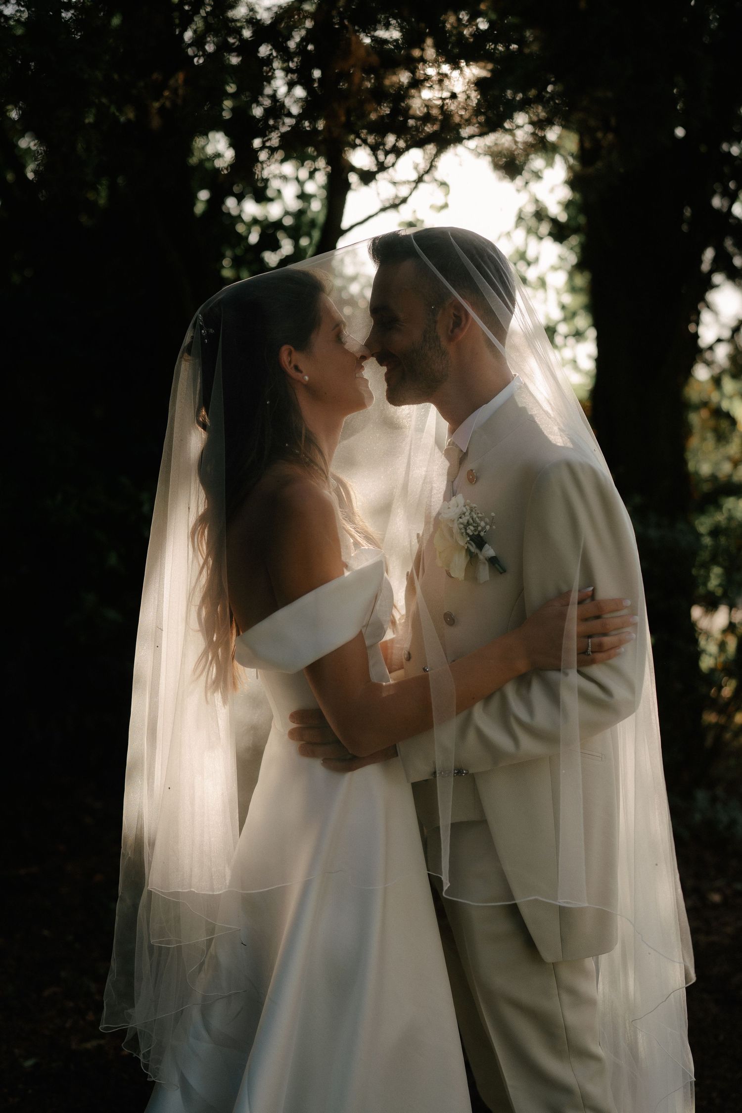Bride and groom share romantic moment under veil in golden backlit forest setting during outdoor wedding ceremony.