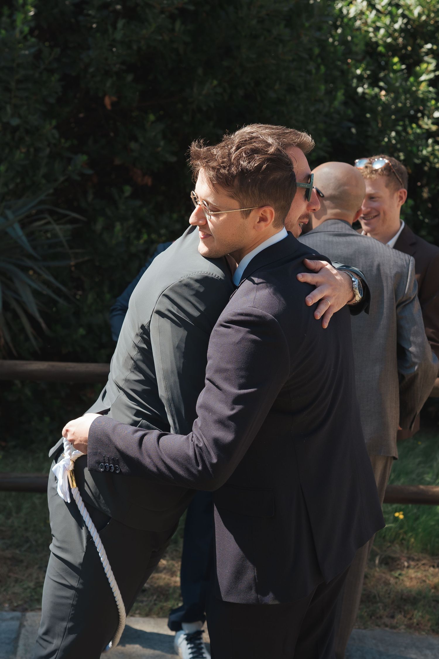 Men in suits embracing at an outdoor gathering, with greenery and smiling guests visible in the background.