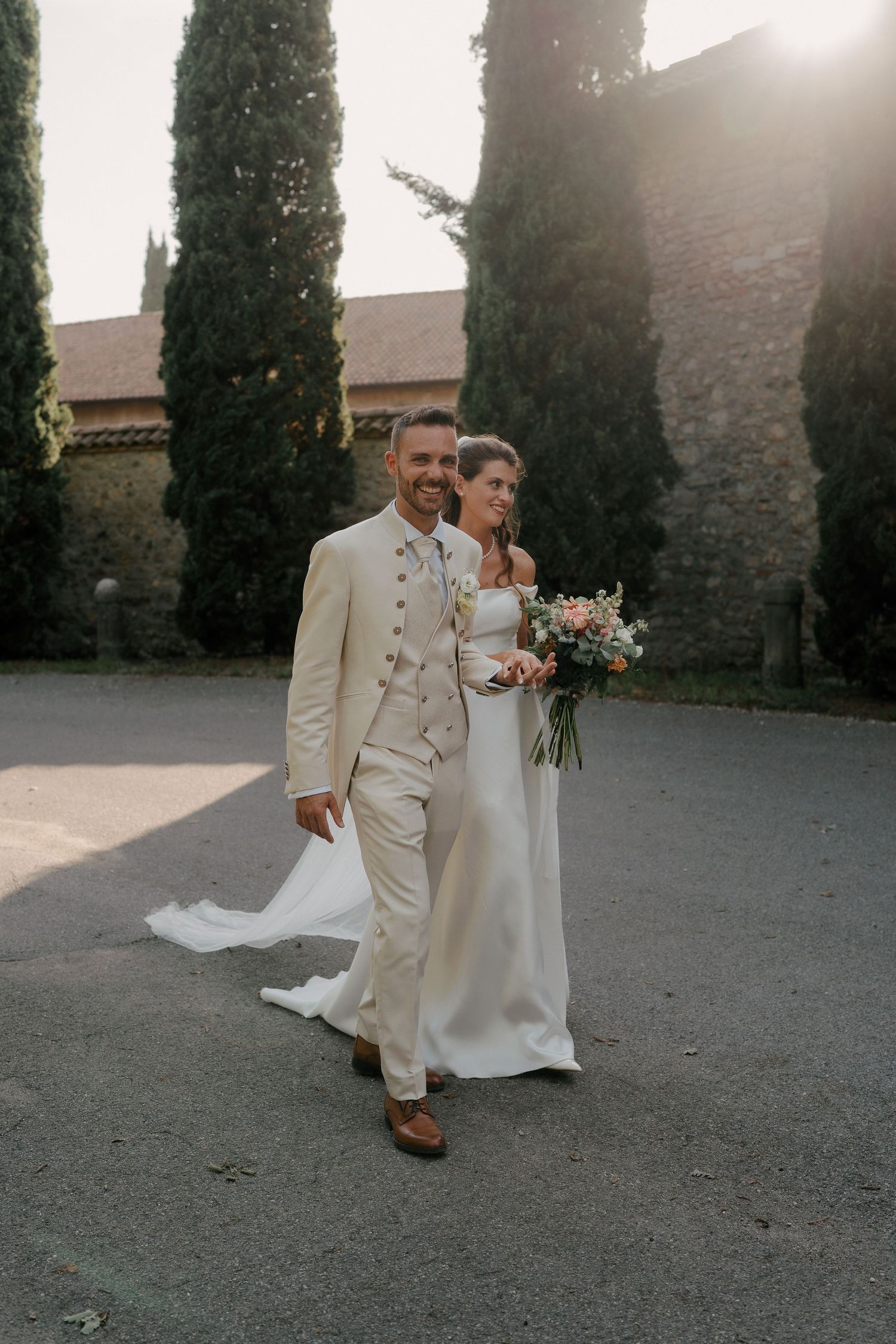 Bride and groom walking outdoors in elegant wedding attire, surrounded by cypress trees and golden sunlight.