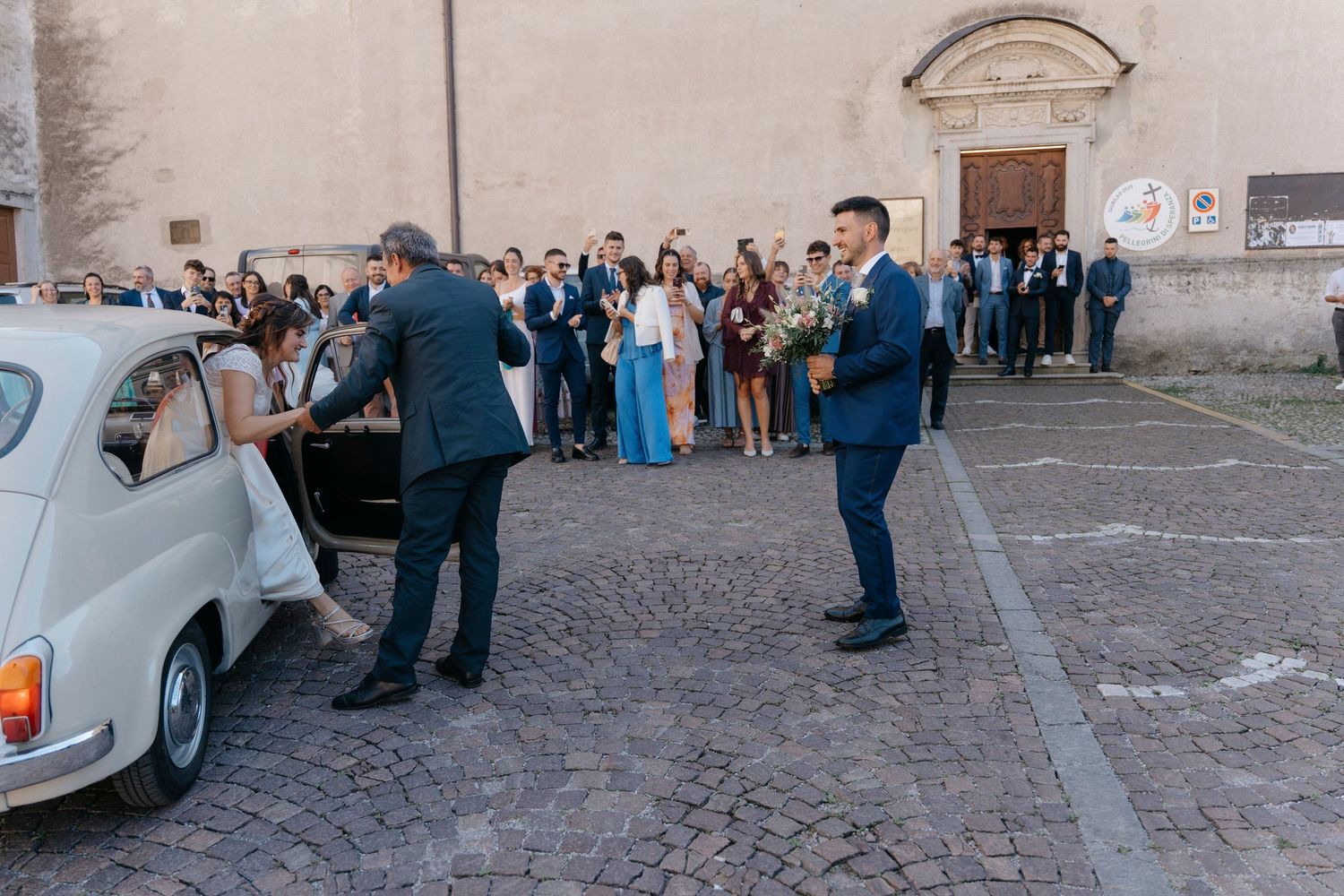 Groom in navy suit waiting outside a church as guests gather near a vintage white car.