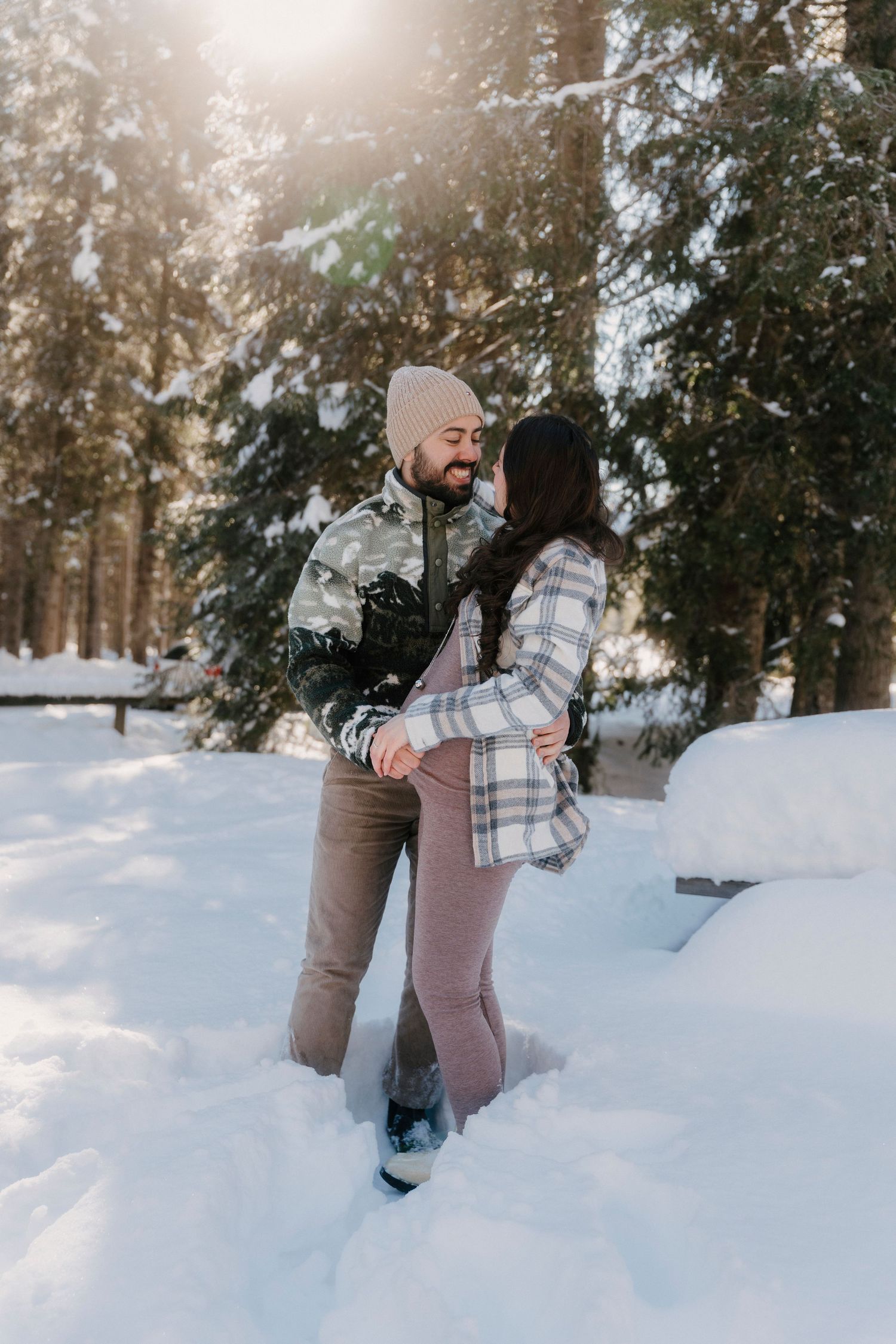 Couple dancing and laughing together in a snowy pine forest with golden sunlight.