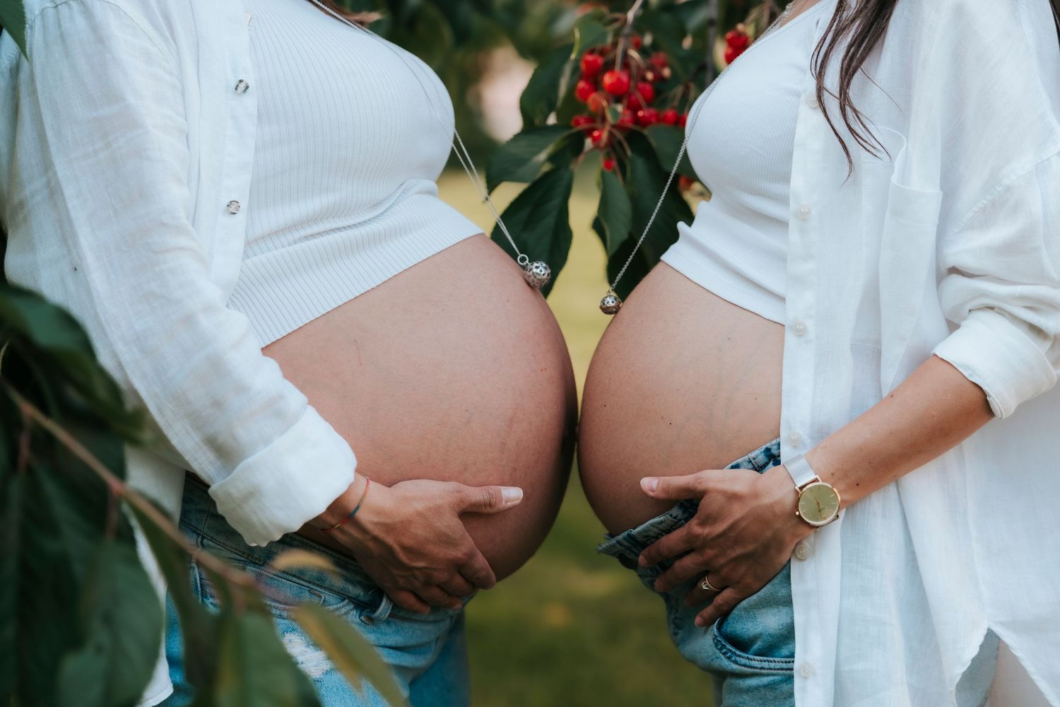 Close-up of two pregnant women in white shirts touching their baby bumps together outdoors.