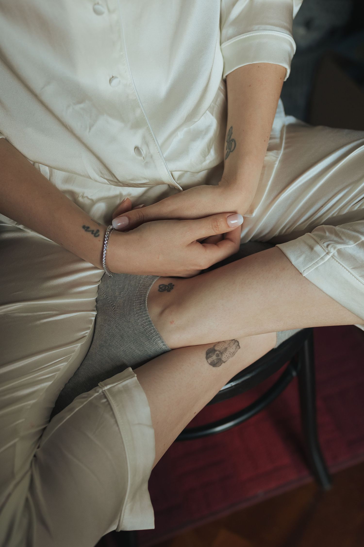 Close-up of bride in white satin robe seated with crossed legs revealing a small ankle tattoo.