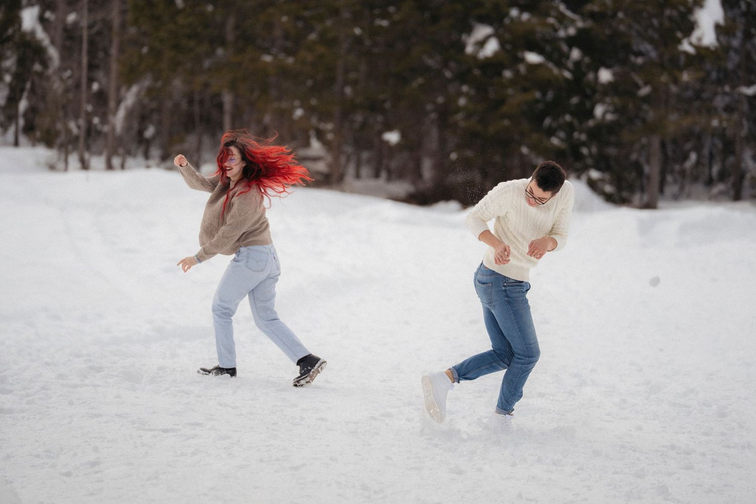 A lively couple plays in the snow, the red-haired woman spinning with hair flying while the man ducks in a snowy winter forest.