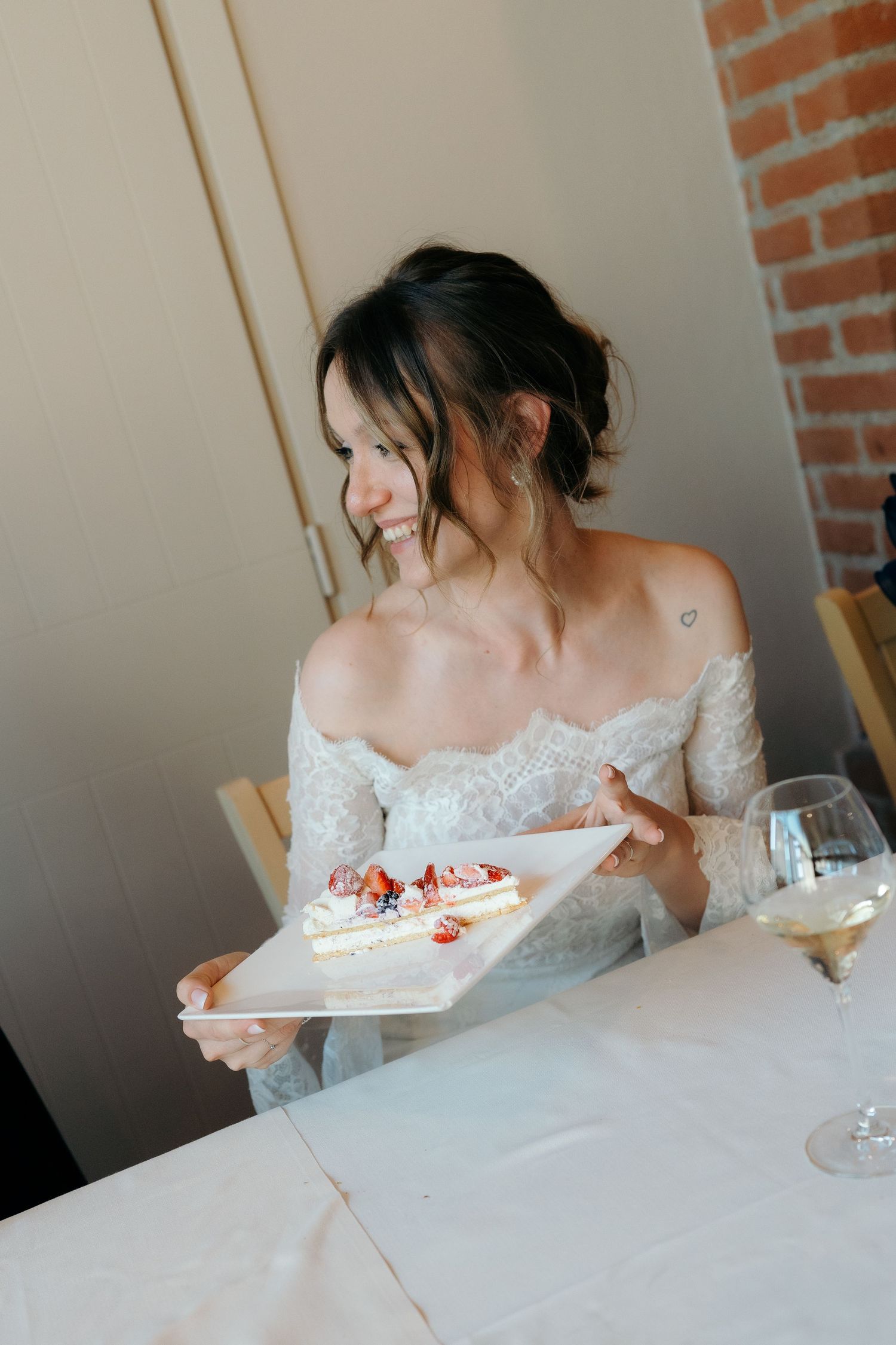 Una sposa felice, con un abito in pizzo con scollo a spalle scoperte, sorride mentre regge un piatto bianco con una fetta di torta nuziale