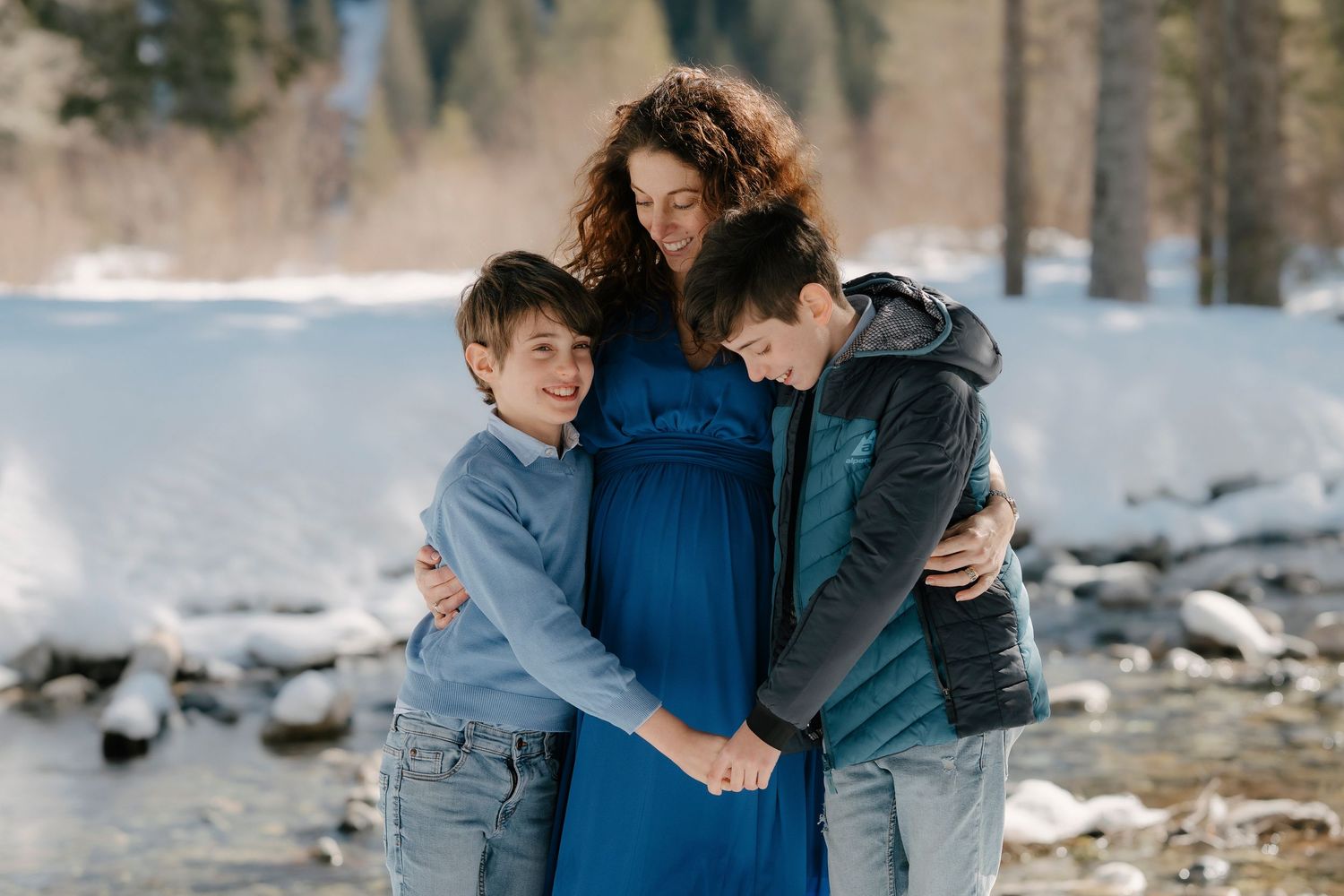 Pregnant woman in blue dress embraced by two boys near snowy winter river.