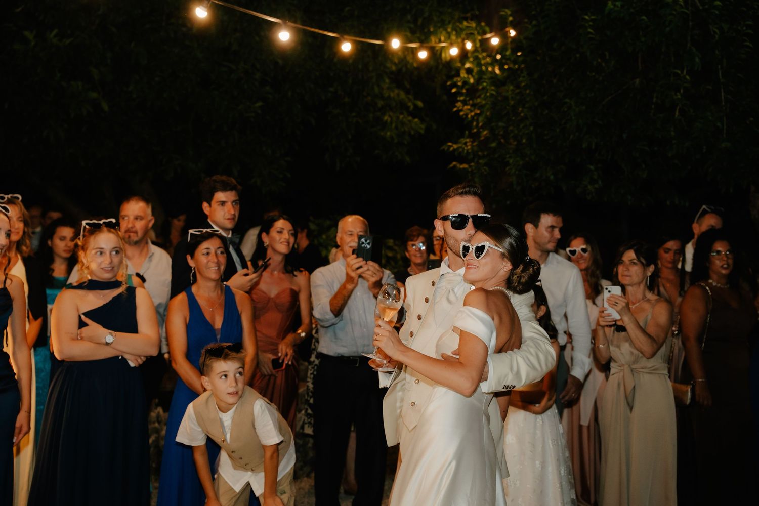 Bride and groom dancing at outdoor wedding reception at night with guests celebrating under string lights.
