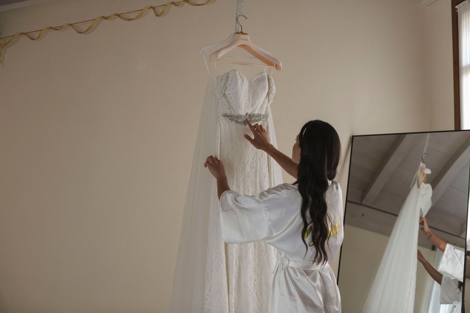 Woman in robe carefully arranging a white wedding dress on a hanger in a bright dressing room.