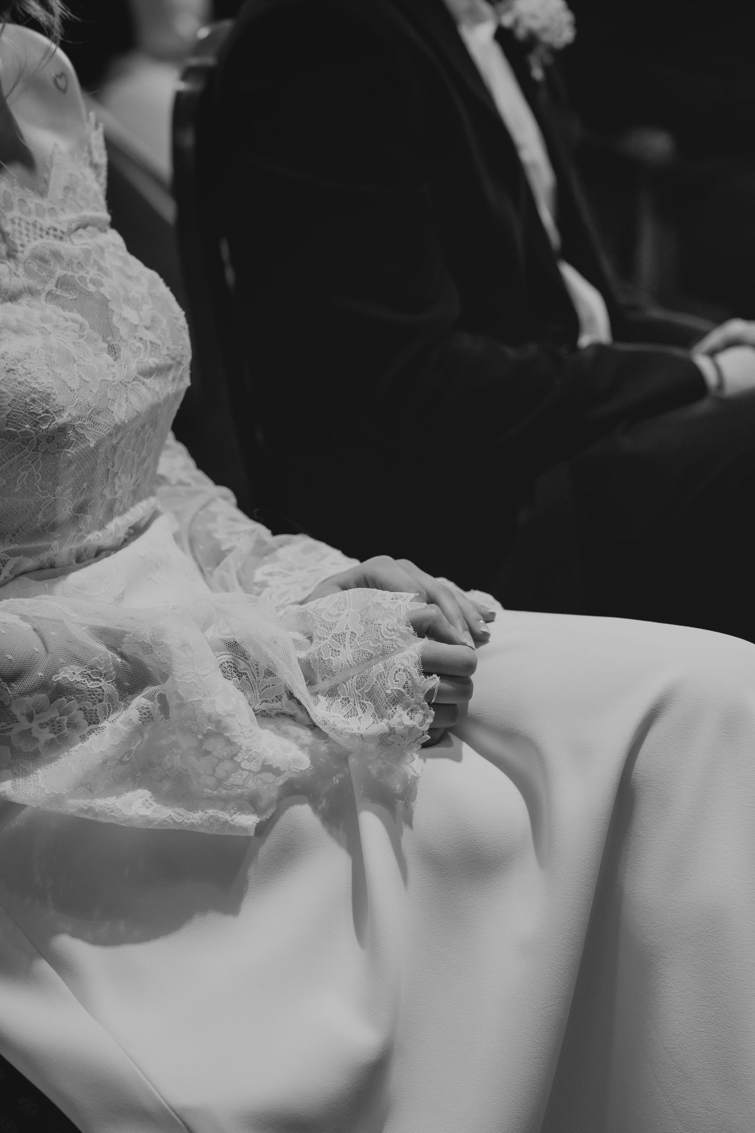 Black and white close-up of bride in lace wedding dress with hands folded in lap during ceremony.
