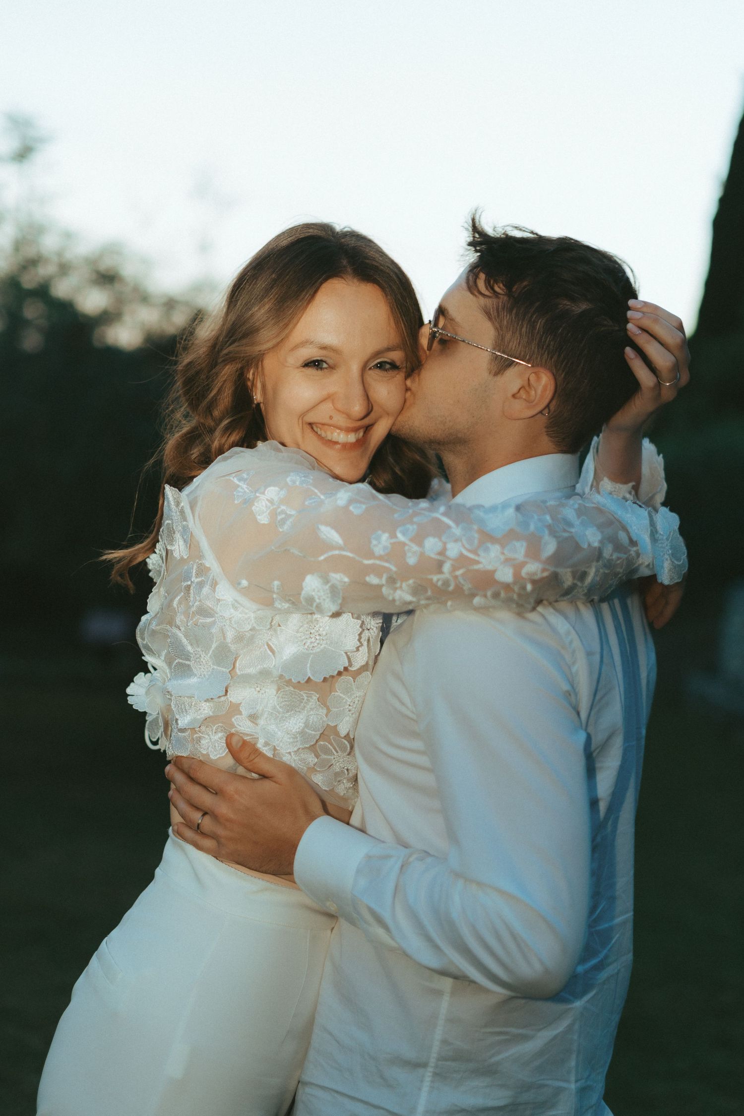 Groom in white shirt kisses smiling bride outdoors at golden hour in lush garden setting.