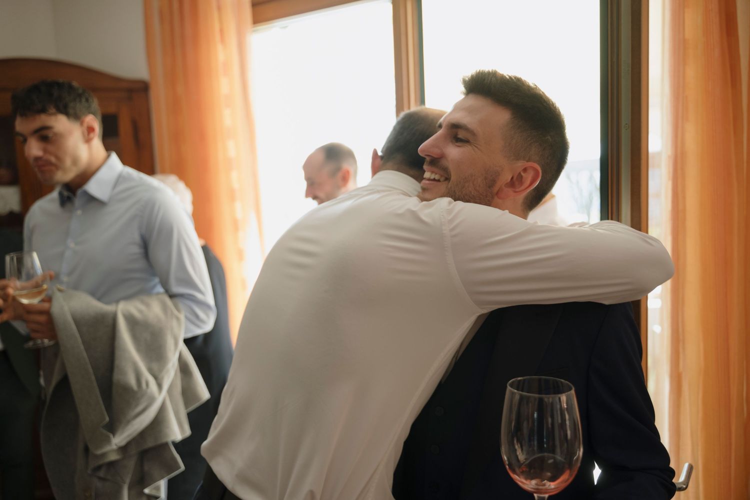 Groom in navy suit sharing a warm embrace with a guest at a pre-wedding gathering indoors.