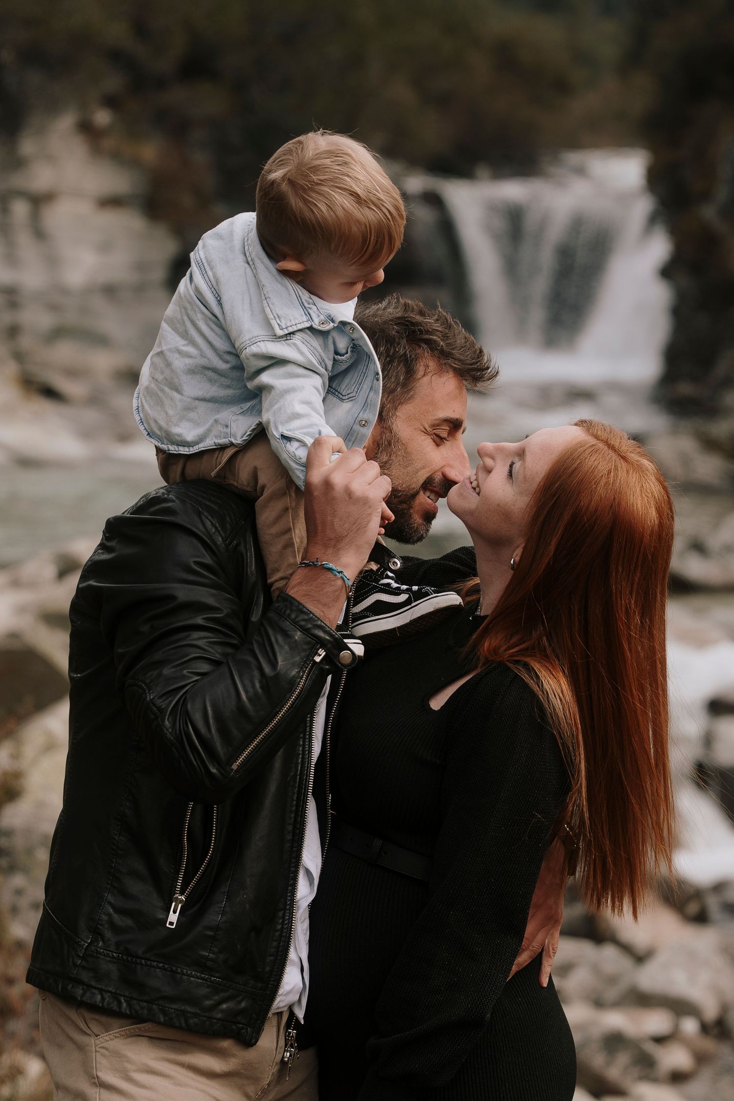 Happy family with redhead mother, bearded father, and toddler posing near a waterfall.