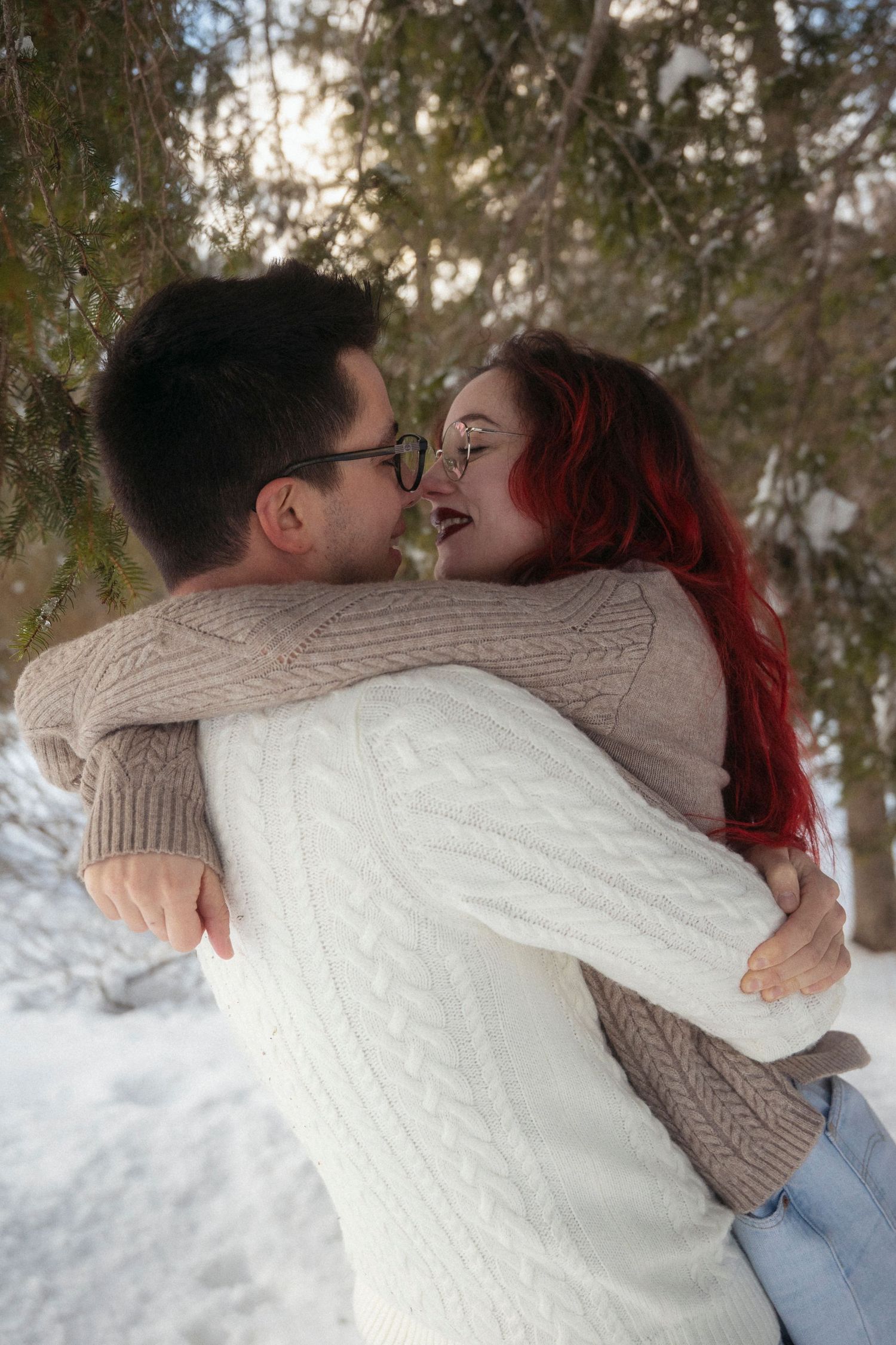 A couple embracing and laughing in a snowy winter park, woman with red hair in cozy sweaters.