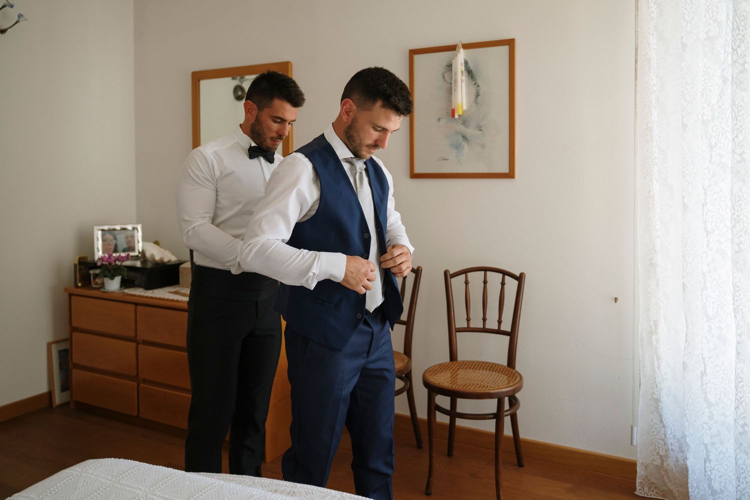 Groom being helped by groomsman to button his navy blue vest while getting ready for wedding.