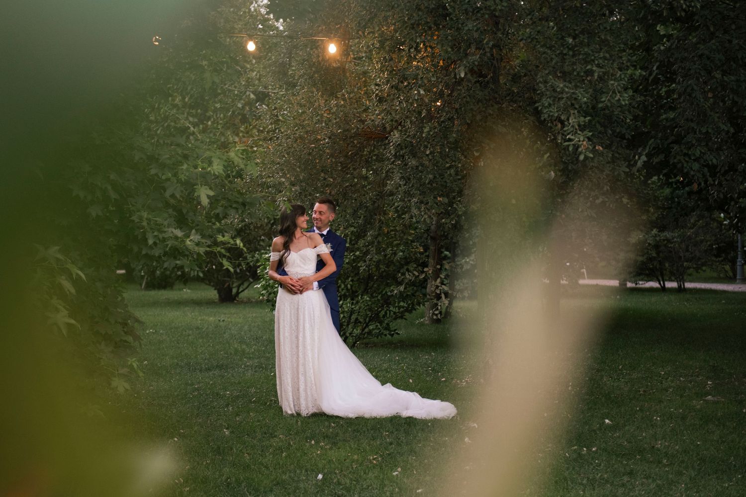Bride and groom embracing in a lush green garden with warm bokeh lights during golden hour wedding photos.