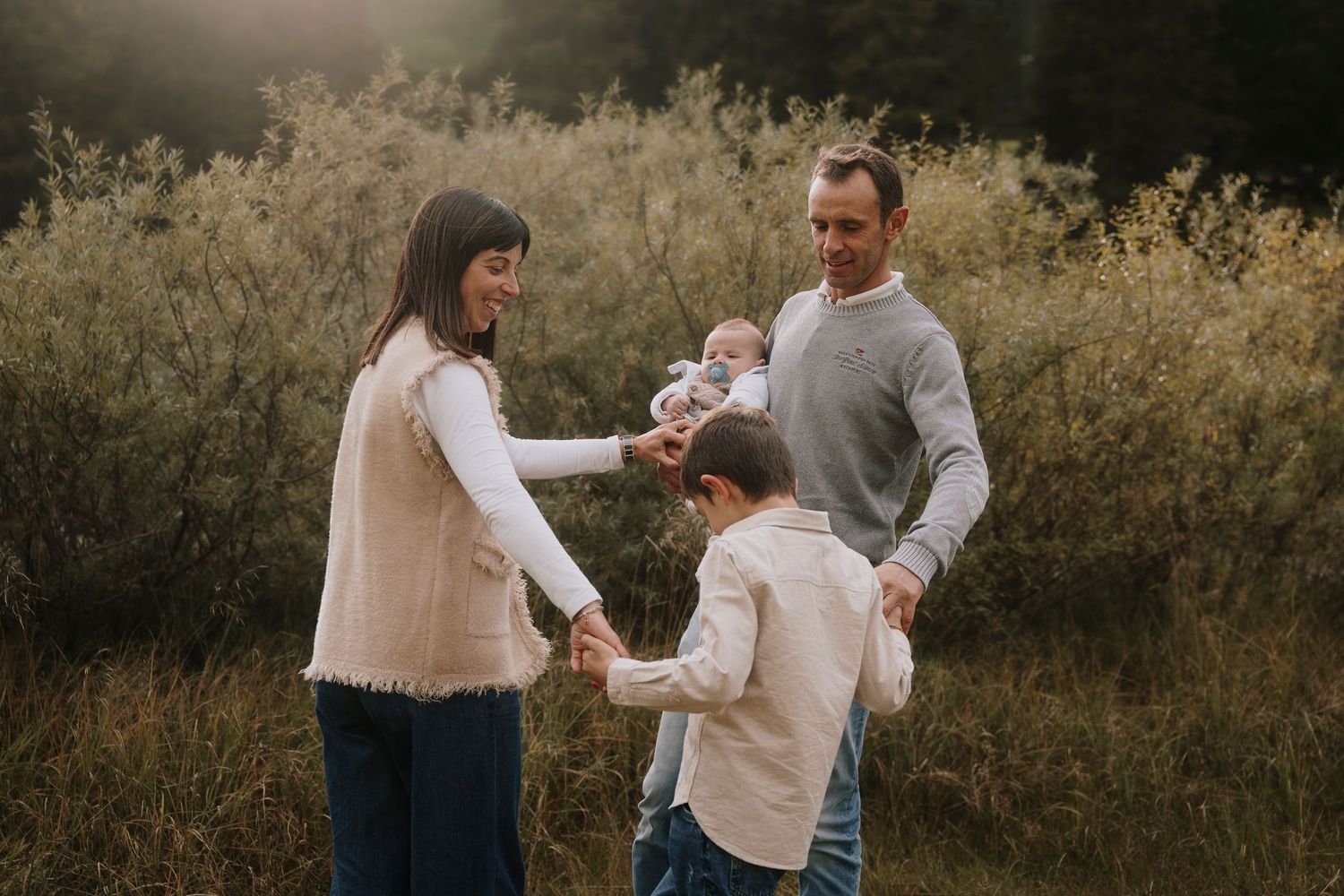 Family of four holding hands together in a golden meadow with lush greenery behind them.