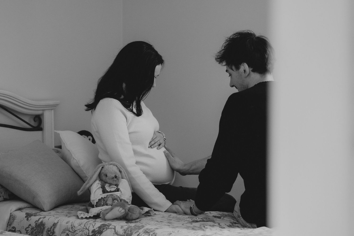Black and white photo of pregnant woman sitting on bed with partner touching her baby bump.