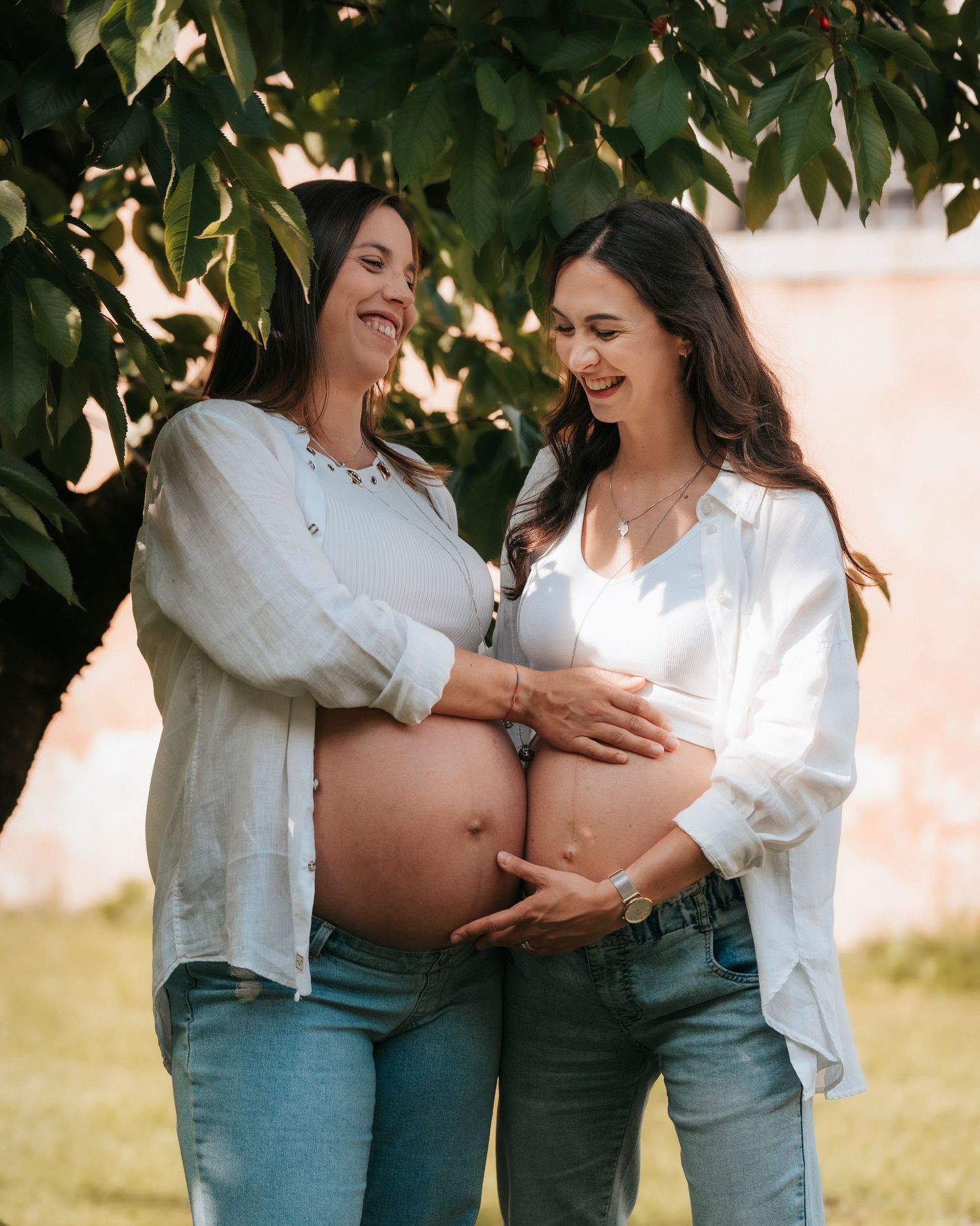 Two pregnant women smiling and touching their baby bumps together under a leafy green tree.
