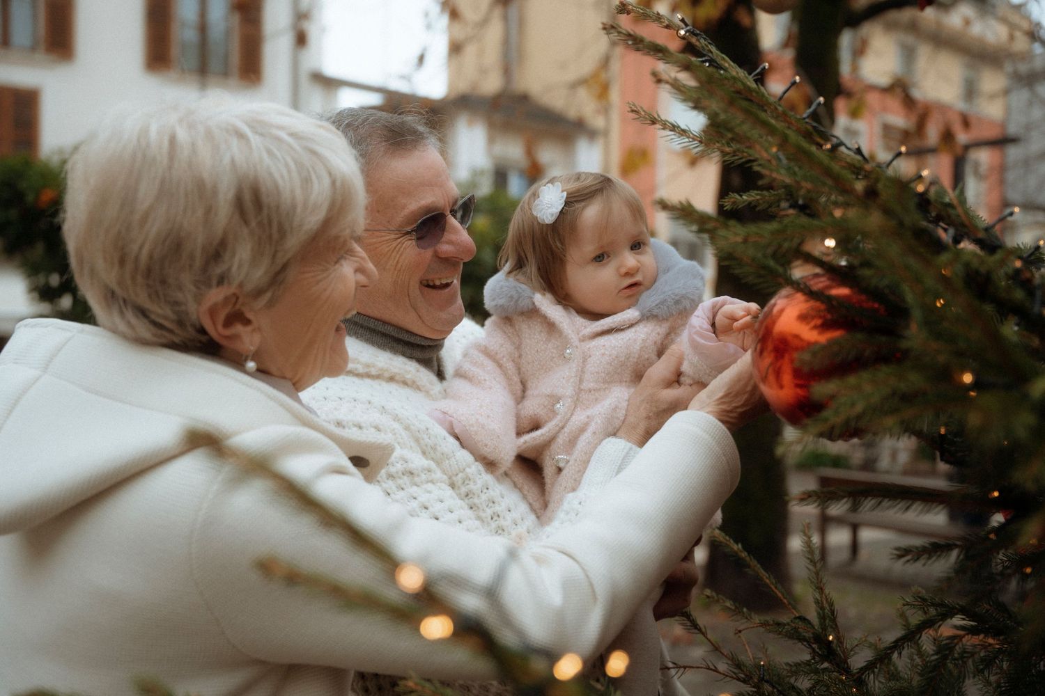 Grandparents and baby girl admiring Christmas tree ornaments outdoors during holiday season.