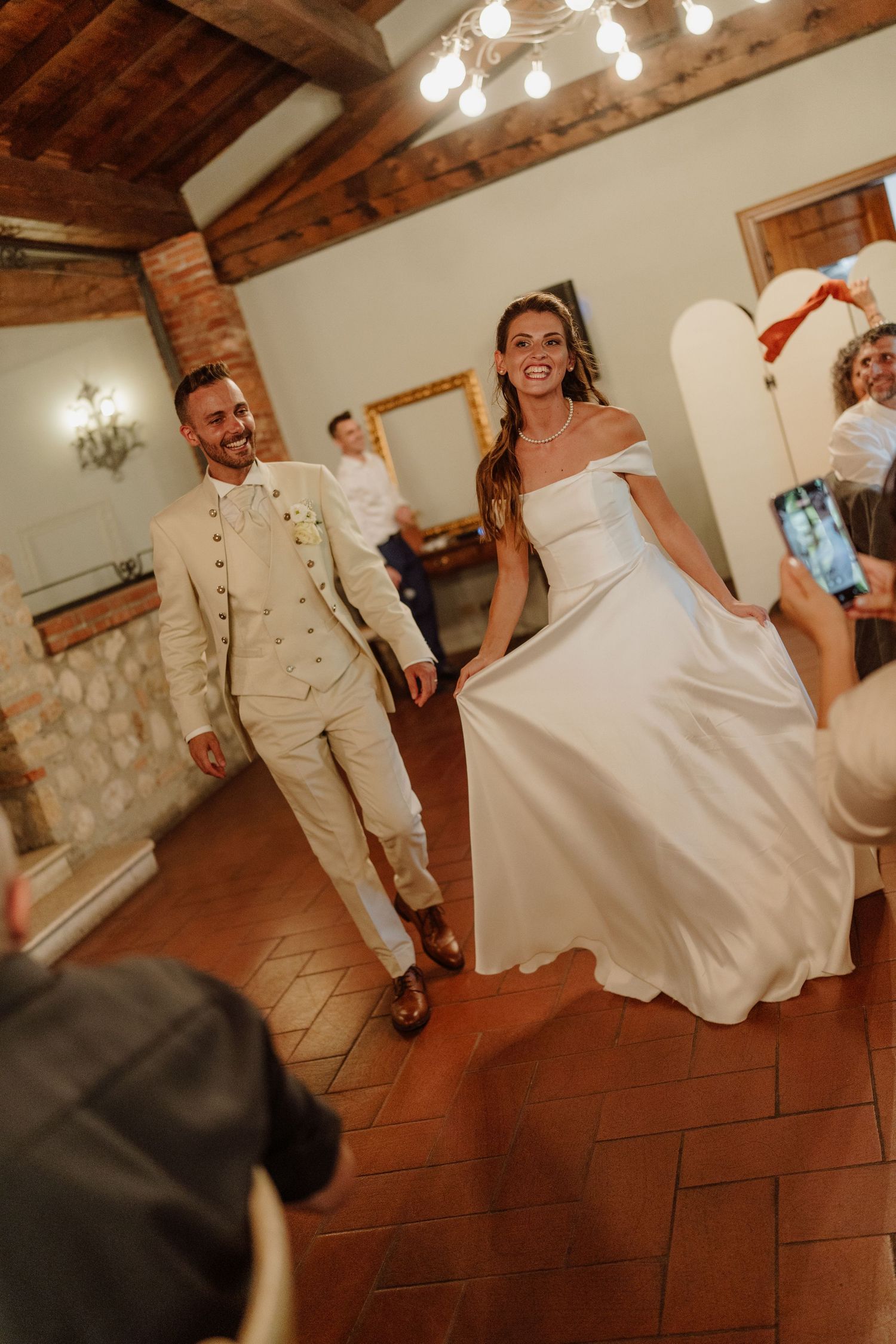 Smiling bride in white off-shoulder gown and groom in beige suit share first dance at elegant rustic wedding venue.