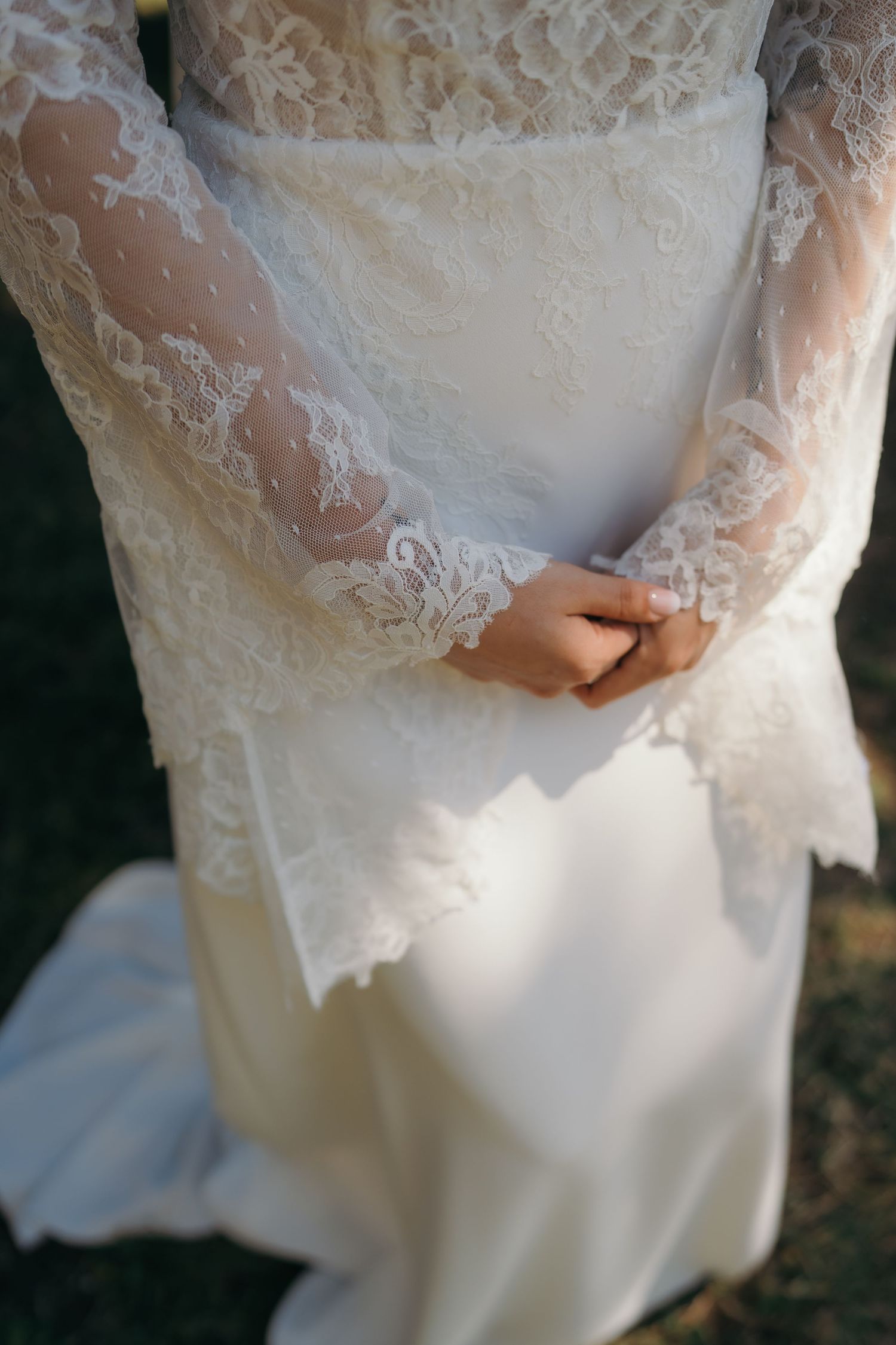 Close-up of a bride's hands clasped together wearing an elegant white lace long-sleeve wedding gown.