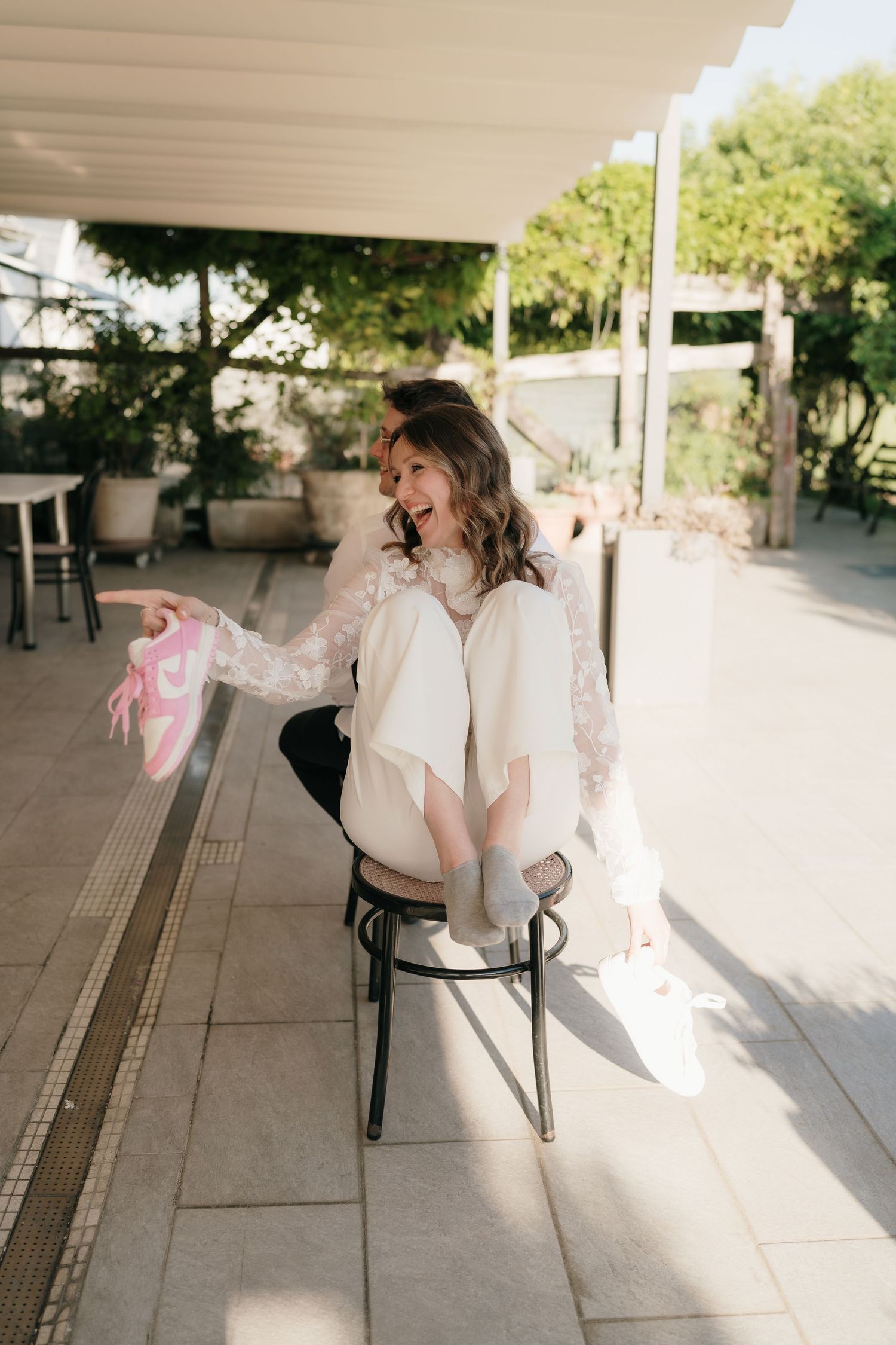 Sposa sorridente con abito in pizzo, seduta su una sedia all'aperto, tiene in mano delle sneakers Nike rosa su una terrazza soleggiata.