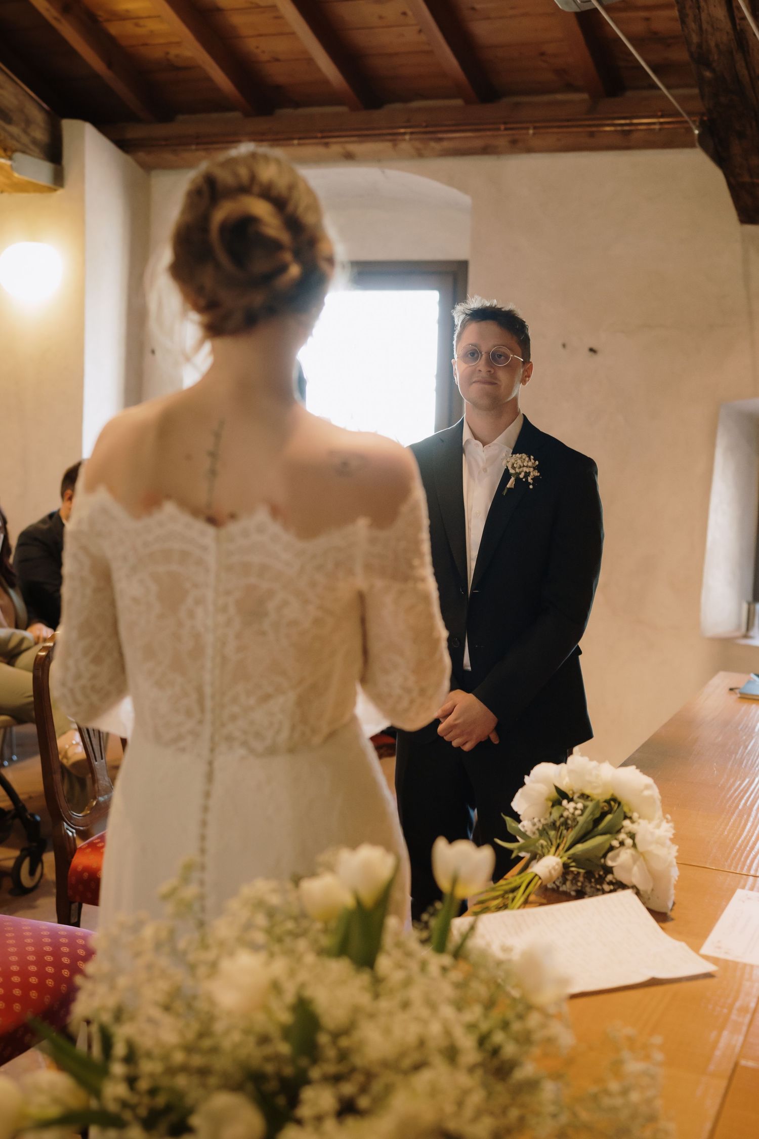 Groom in black suit smiling at bride in lace wedding dress during indoor ceremony with white tulips.