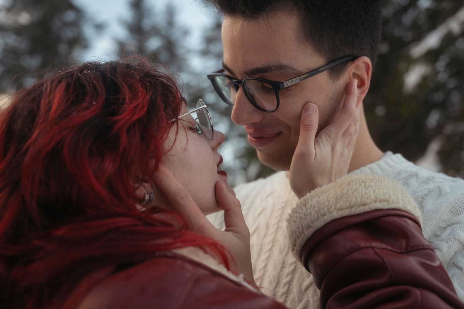 A romantic couple nearly kissing in a snowy setting, the woman with red hair gently touching the mans face while both wear glasses.