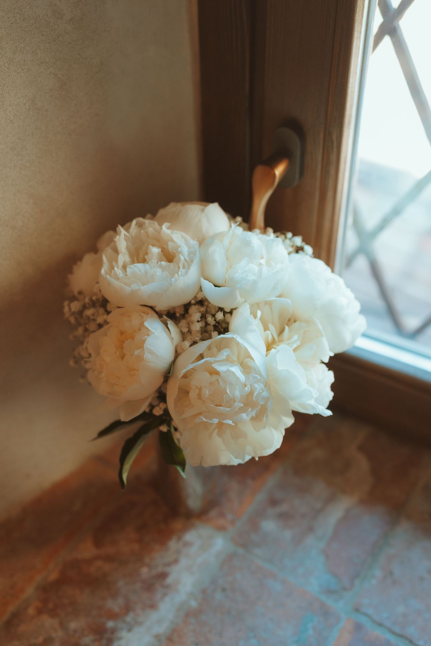 Bridal bouquet of white peonies and baby's breath in a vase beside a sunlit lattice window.