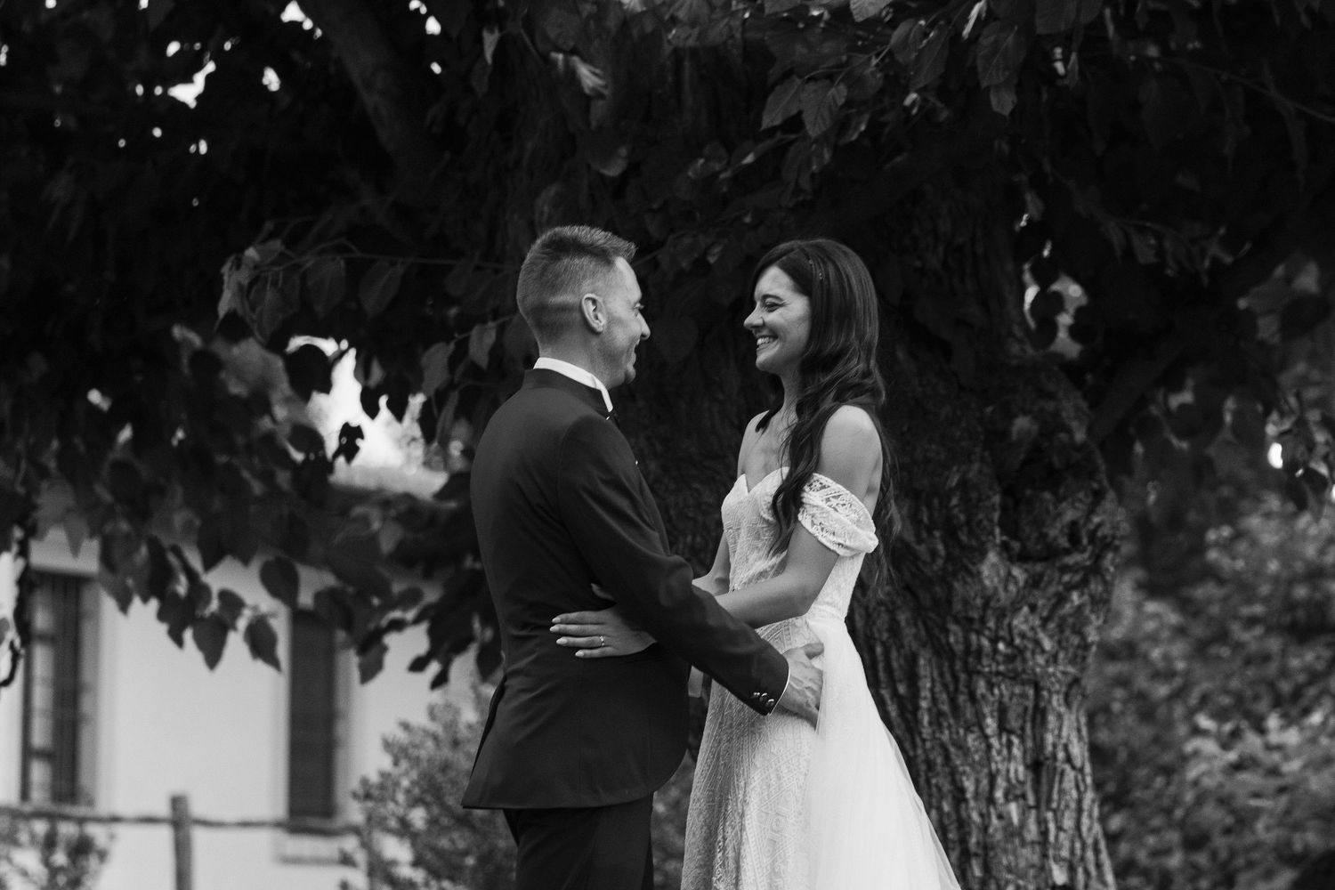Black and white photo of a couple holding hands and smiling under a tree in wedding attire.