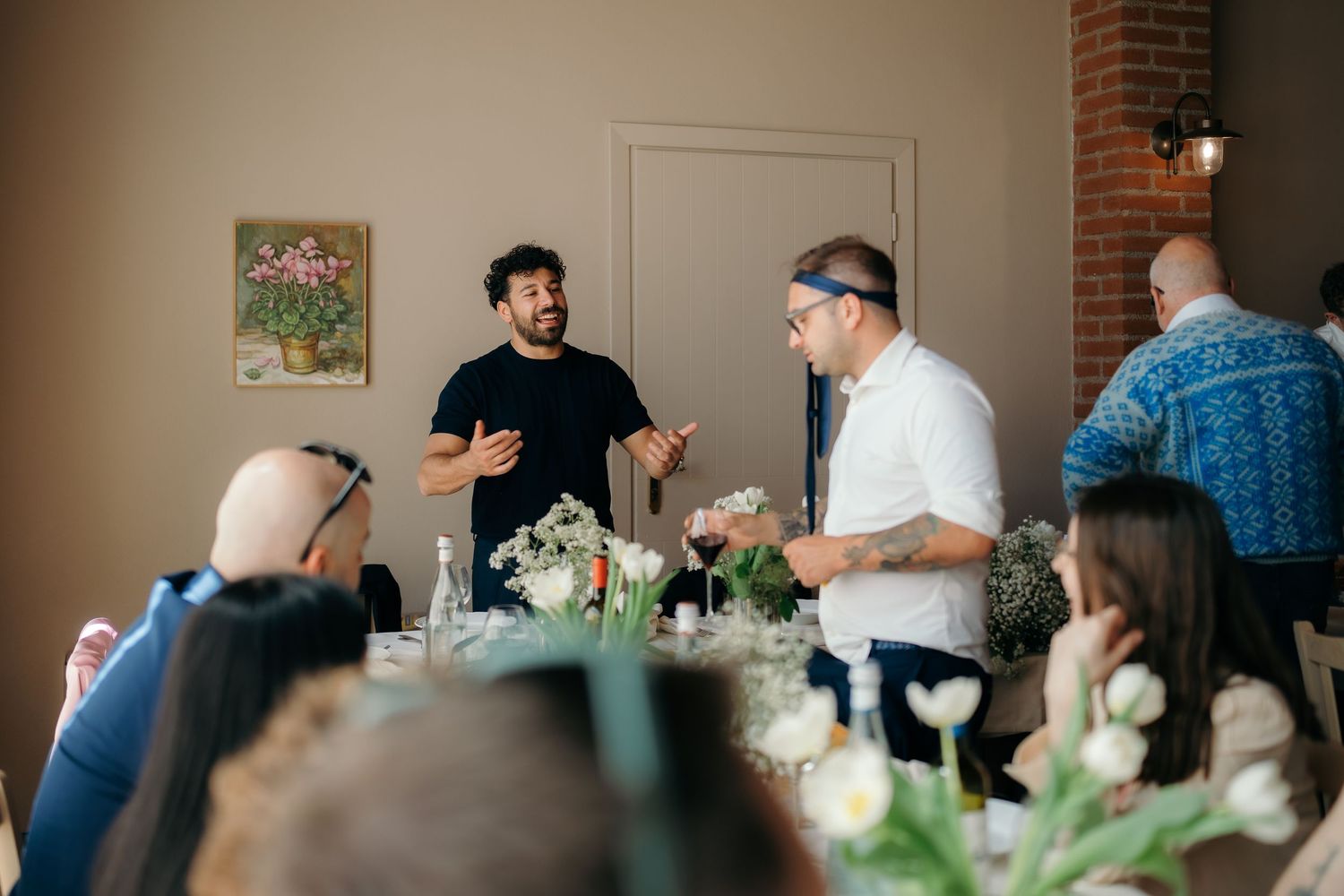 Two men conversing at an elegant indoor dinner event with floral centerpieces and seated guests nearby.