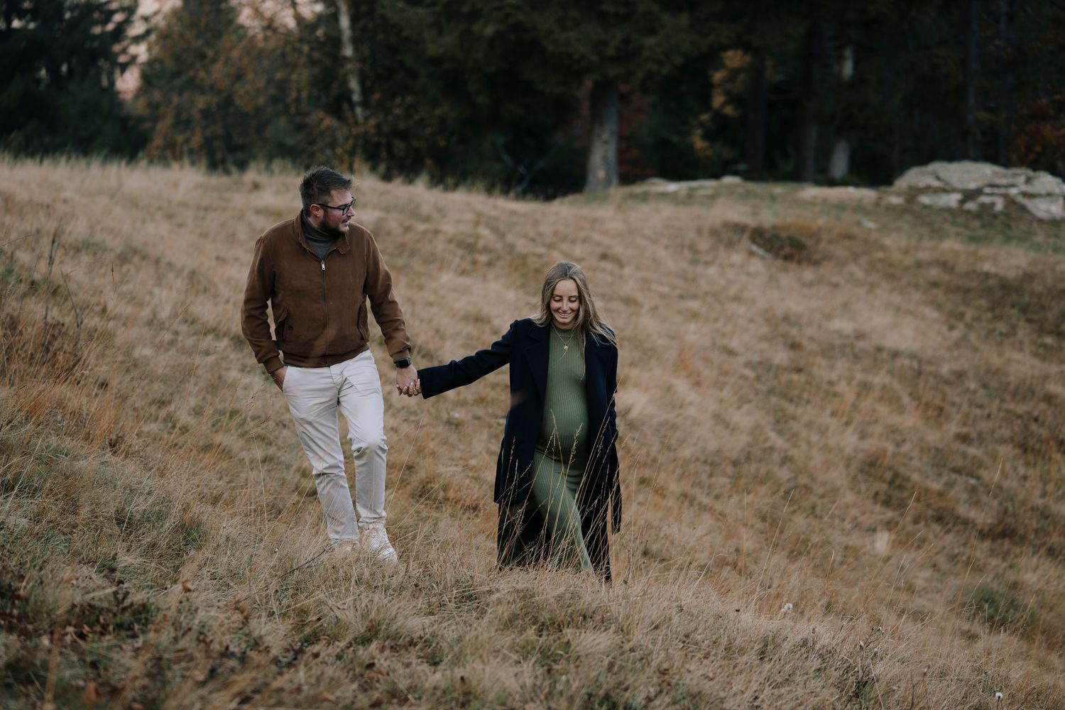 Couple holding hands walking through golden autumn meadow with trees in background.