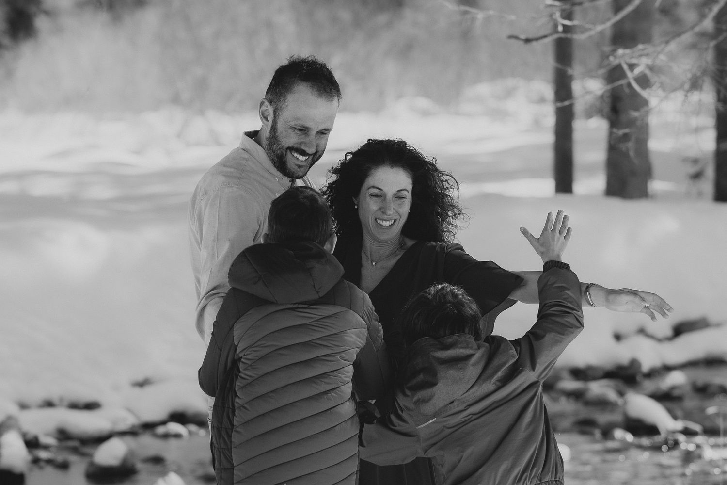 A happy family shares a warm embrace outdoors in a snowy winter landscape, captured in black and white.