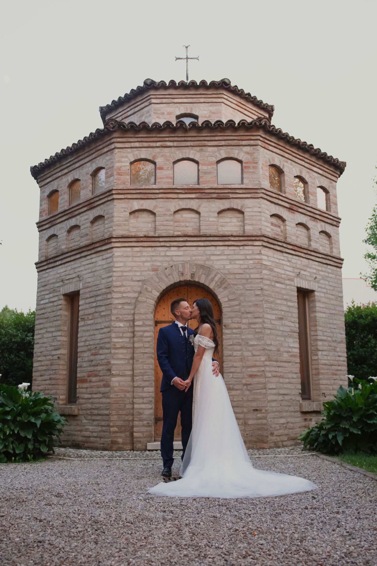 Bride and groom sharing a kiss in front of an octagonal brick chapel with a cross on top.