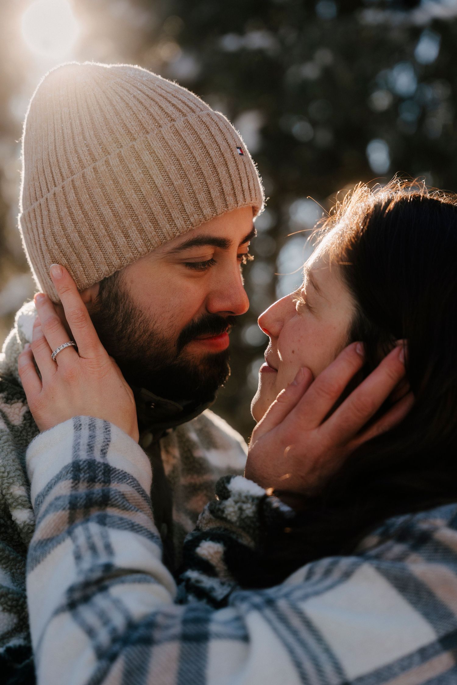 Romantic couple gazing into each other's eyes in a sunlit snowy forest, gently holding each other's faces.