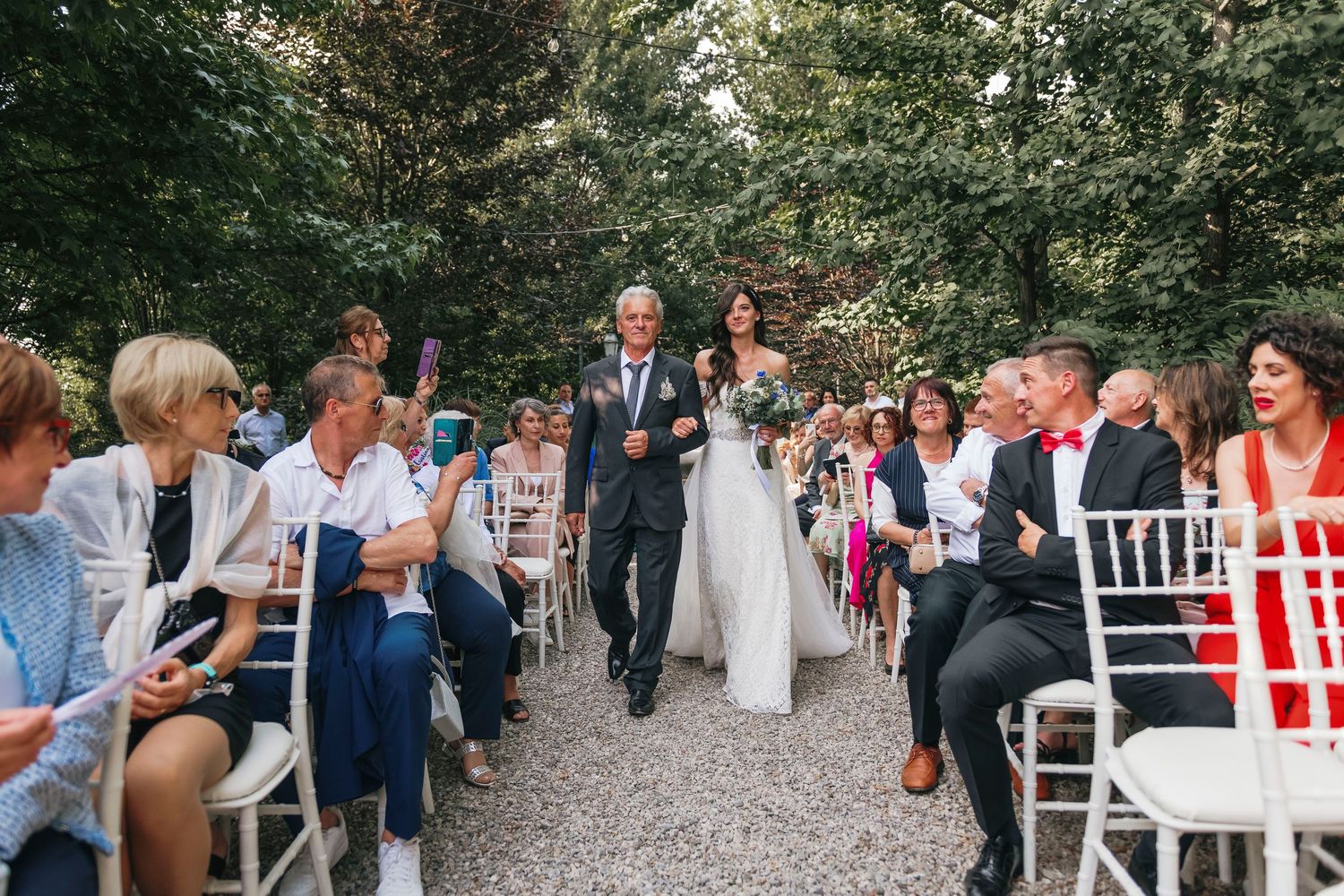 Bride walks down outdoor aisle with escort past seated wedding guests at garden ceremony.