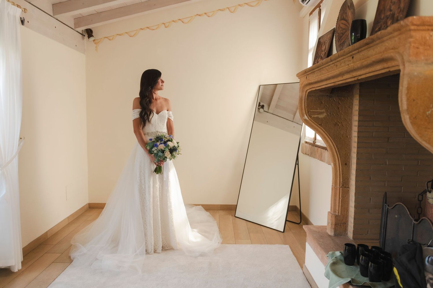 Bride in off-shoulder wedding gown holding bouquet beside mirror in elegant room.