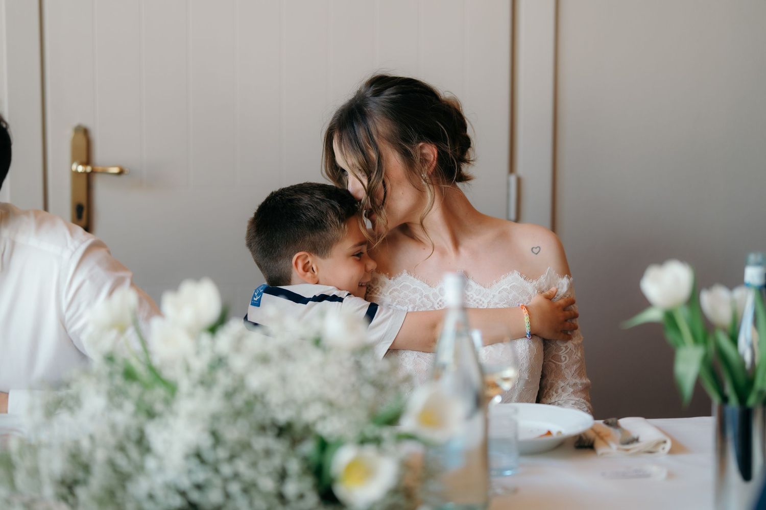 Bride in lace gown sharing tender moment with young boy at elegant wedding reception table with white flowers.