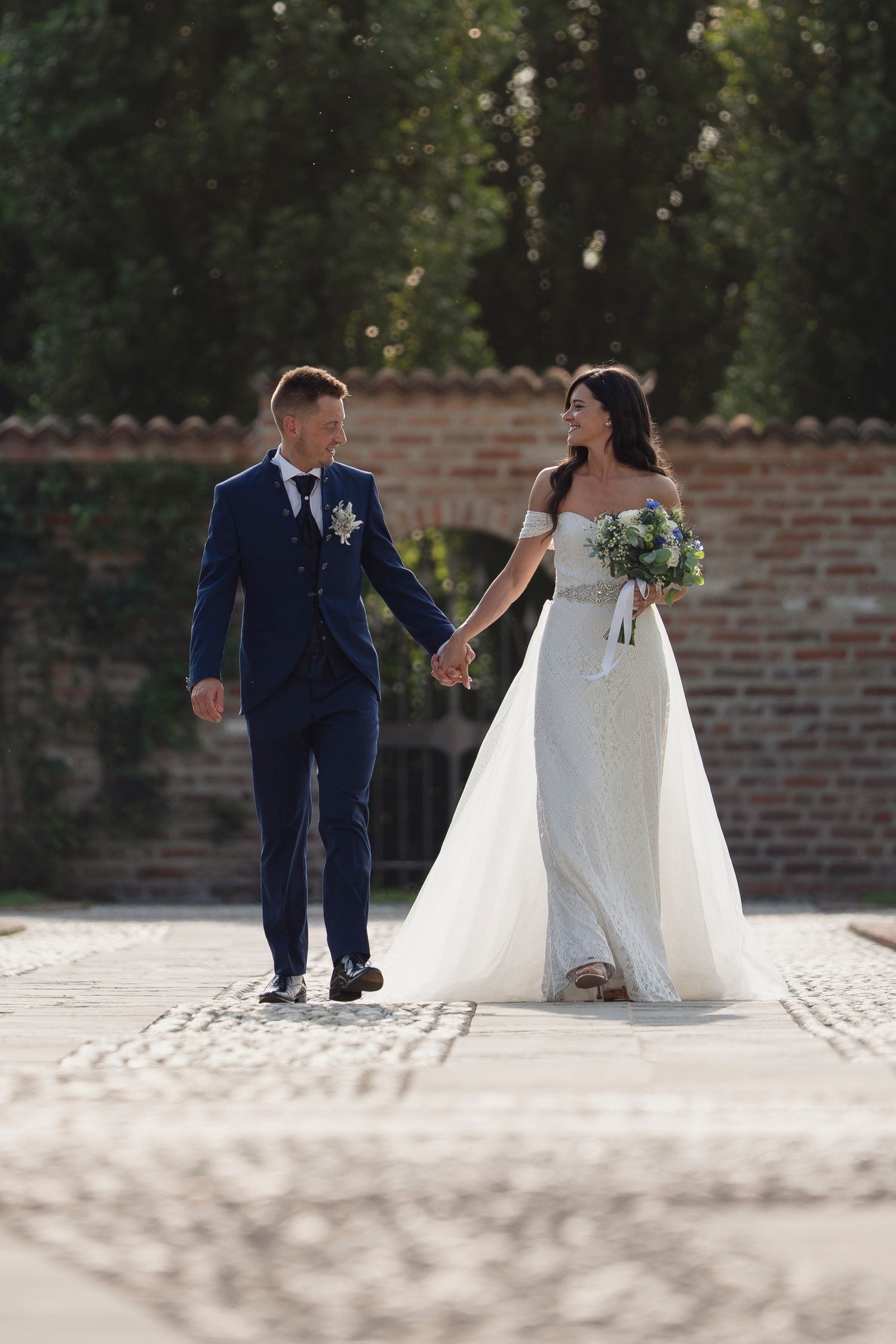 Bride and groom walk hand-in-hand on cobblestone path, she in white gown holding bouquet, he in navy suit.