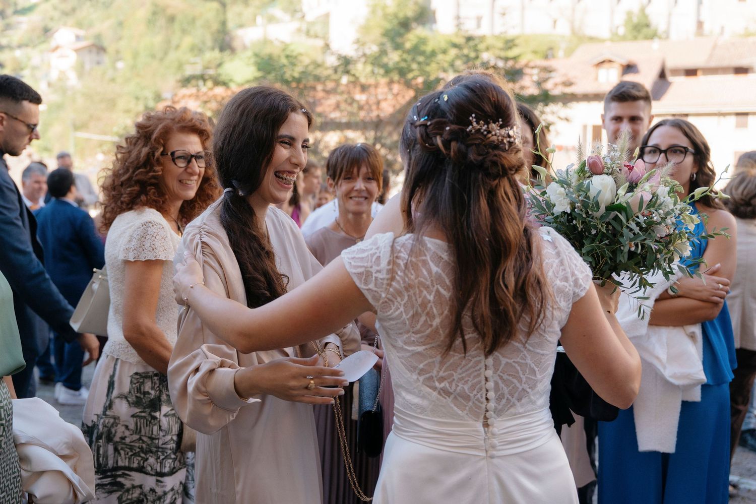 Bride in lace wedding dress receives congratulations from smiling guests while holding a floral bouquet outdoors.