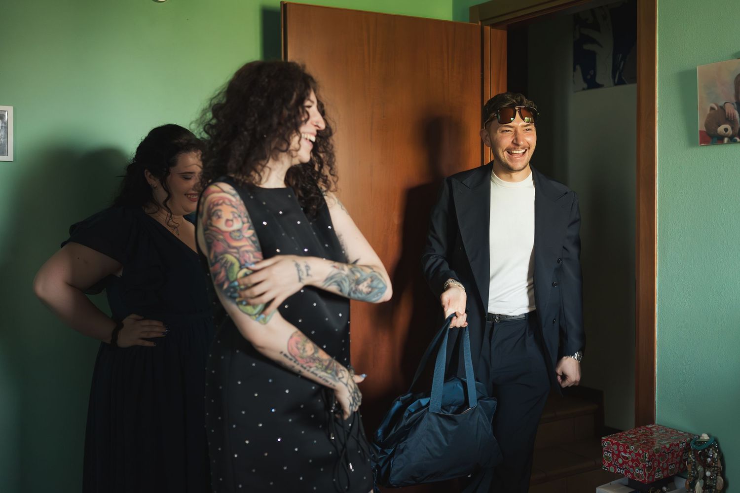 Wedding guests laughing and chatting in a green room while someone carries a suit bag through the door.