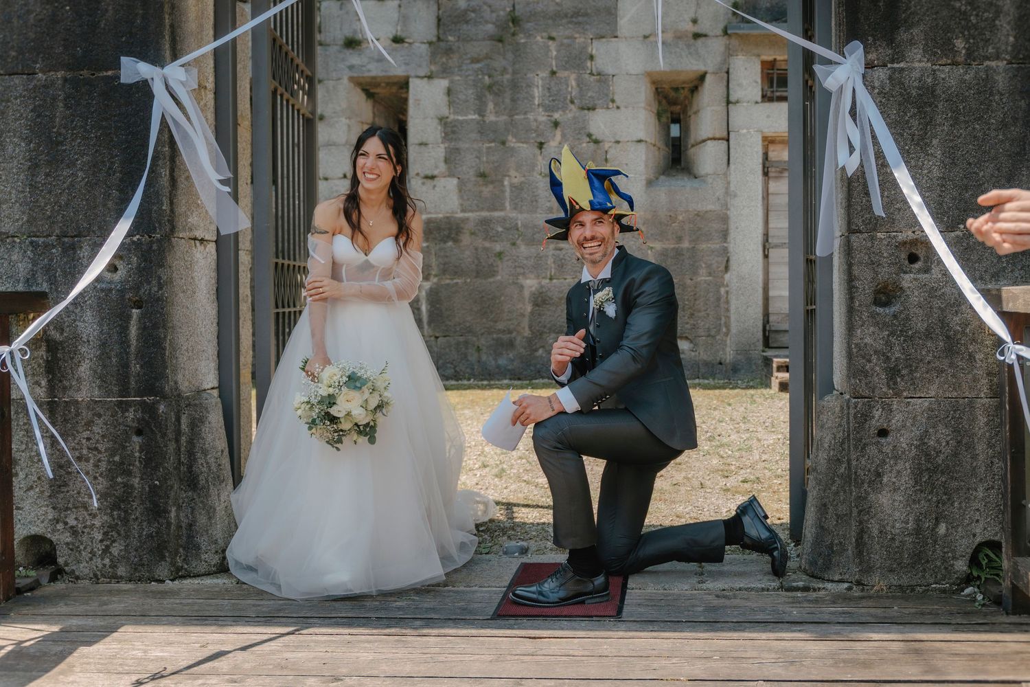 Una sposa e uno sposo in posa giocosa all'ingresso di una storica struttura in pietra, nel giorno del loro matrimonio.