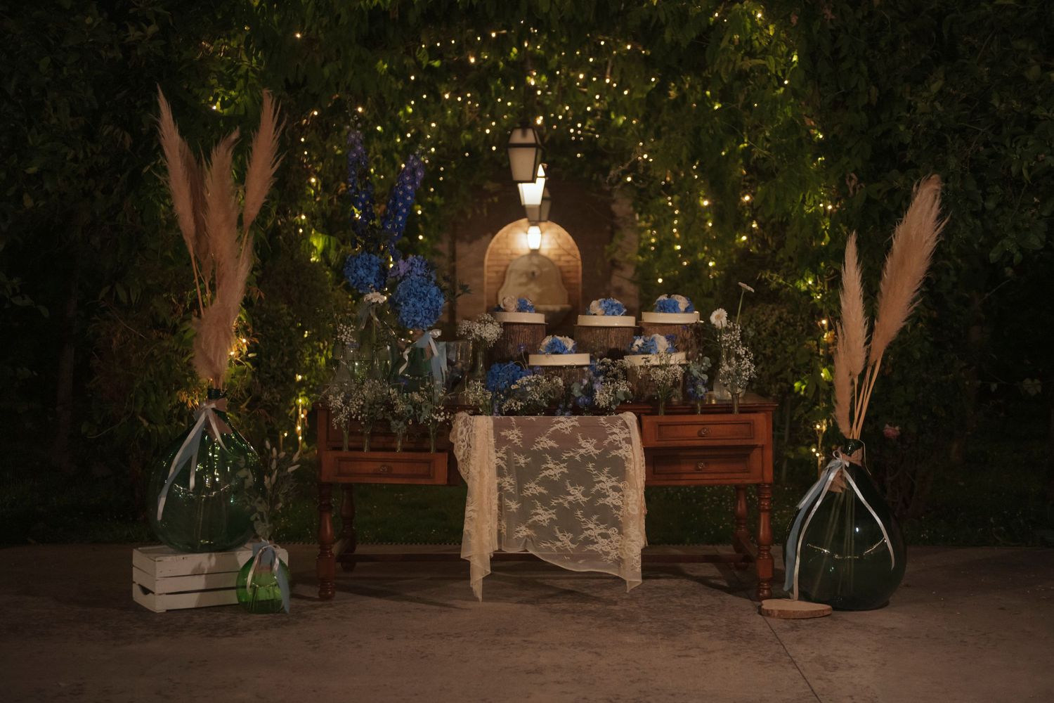 Elegant outdoor dessert table with fairy lights, lace tablecloth, and tall floral arrangements in a garden setting at night.