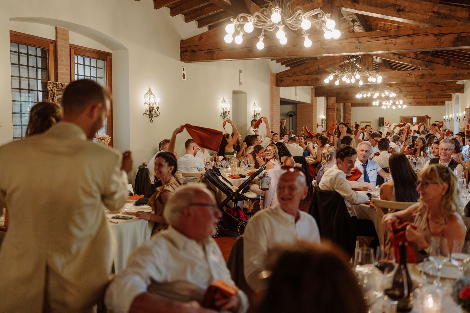 Elegant wedding reception in a rustic venue with exposed beams, guests seated at tables, warm lighting, and a speaker addressing the crowd.