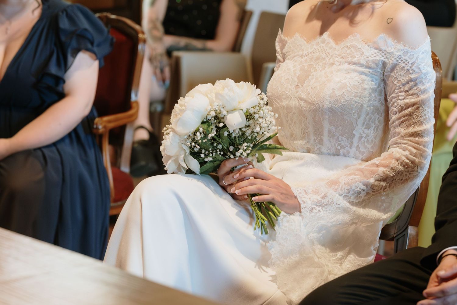 Bride in lace wedding dress holding white rose bouquet with baby's breath while seated at ceremony.