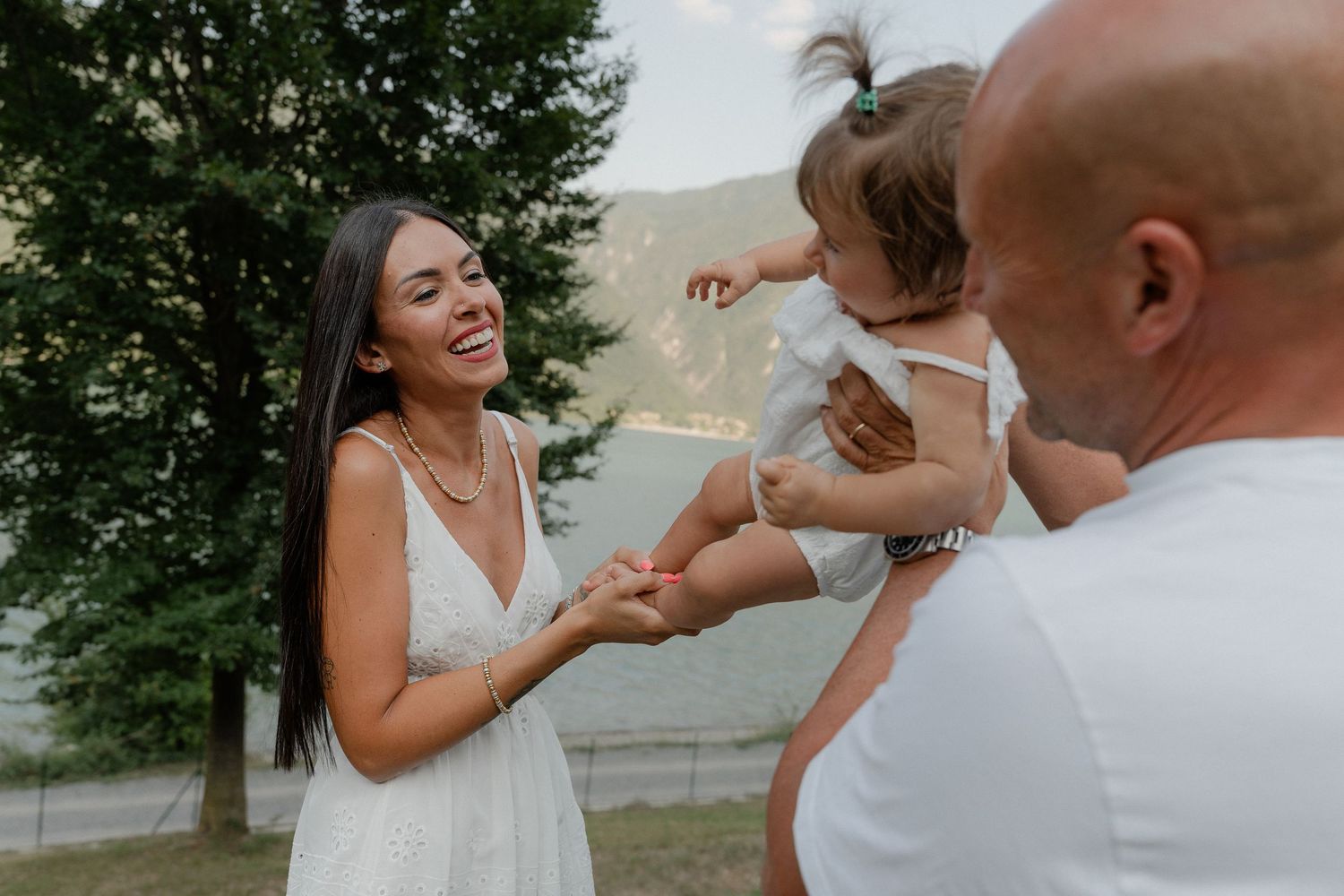 A laughing woman in white dress holds hands with a man lifting their baby girl outdoors near trees.