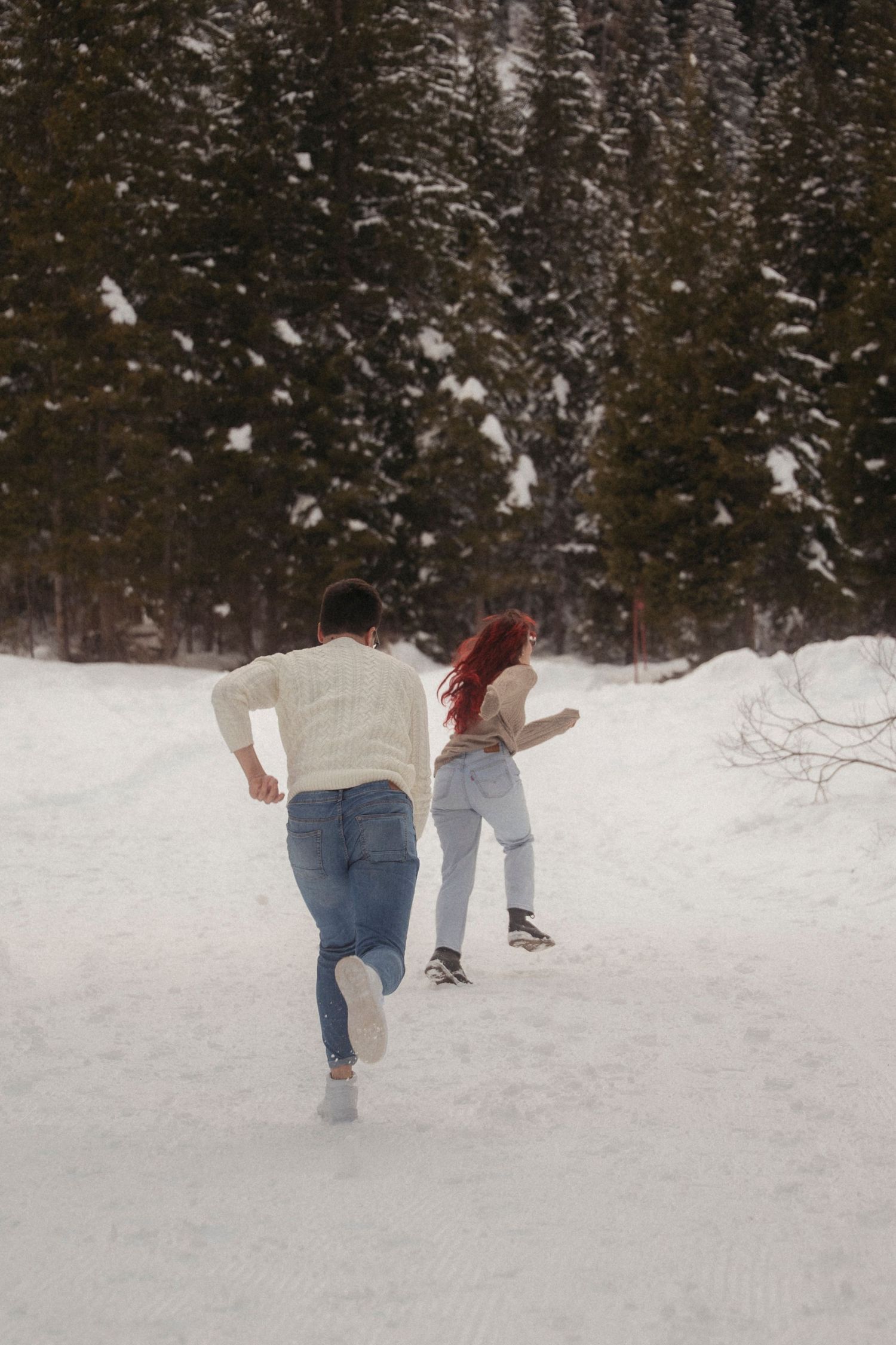 A couple runs playfully through a snowy winter landscape toward a forest of snow-dusted evergreen trees, viewed from behind.