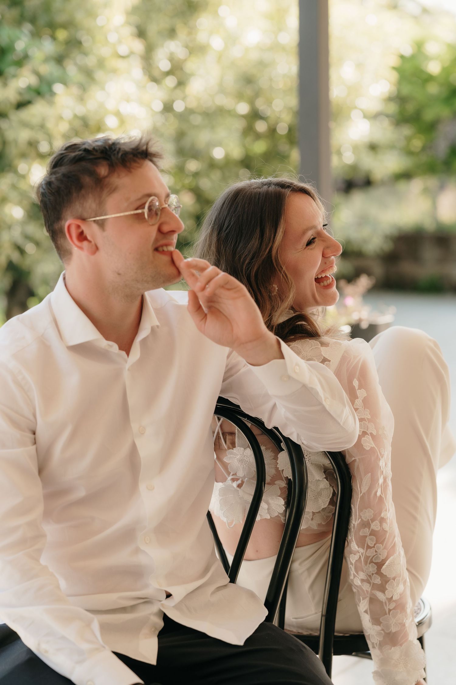 Una sposa sorridente in abito di pizzo tocca scherzosamente le labbra dello sposo mentre siedono insieme ridendo in un giardino