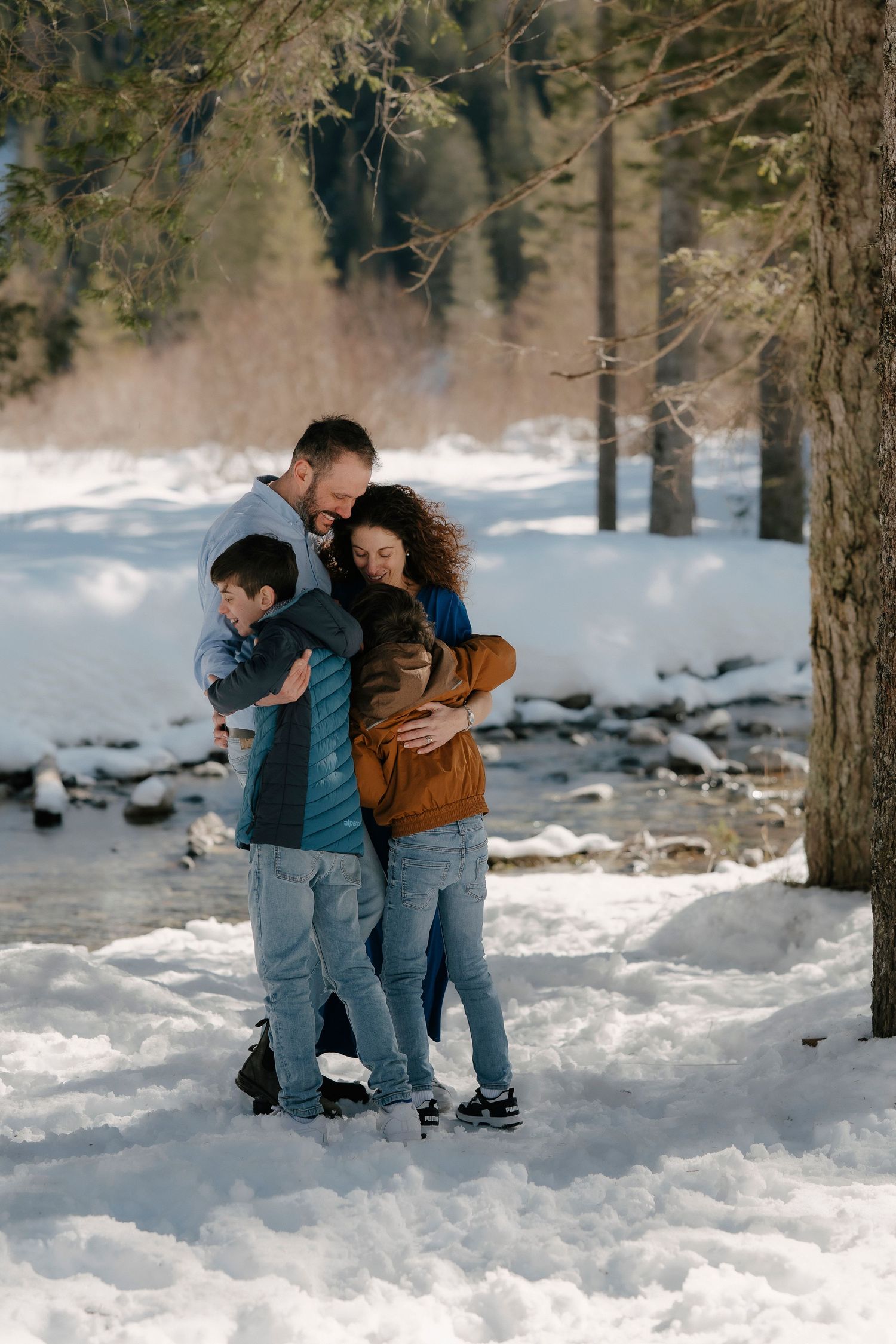 Family of four hugging in snowy winter landscape near a frozen stream.