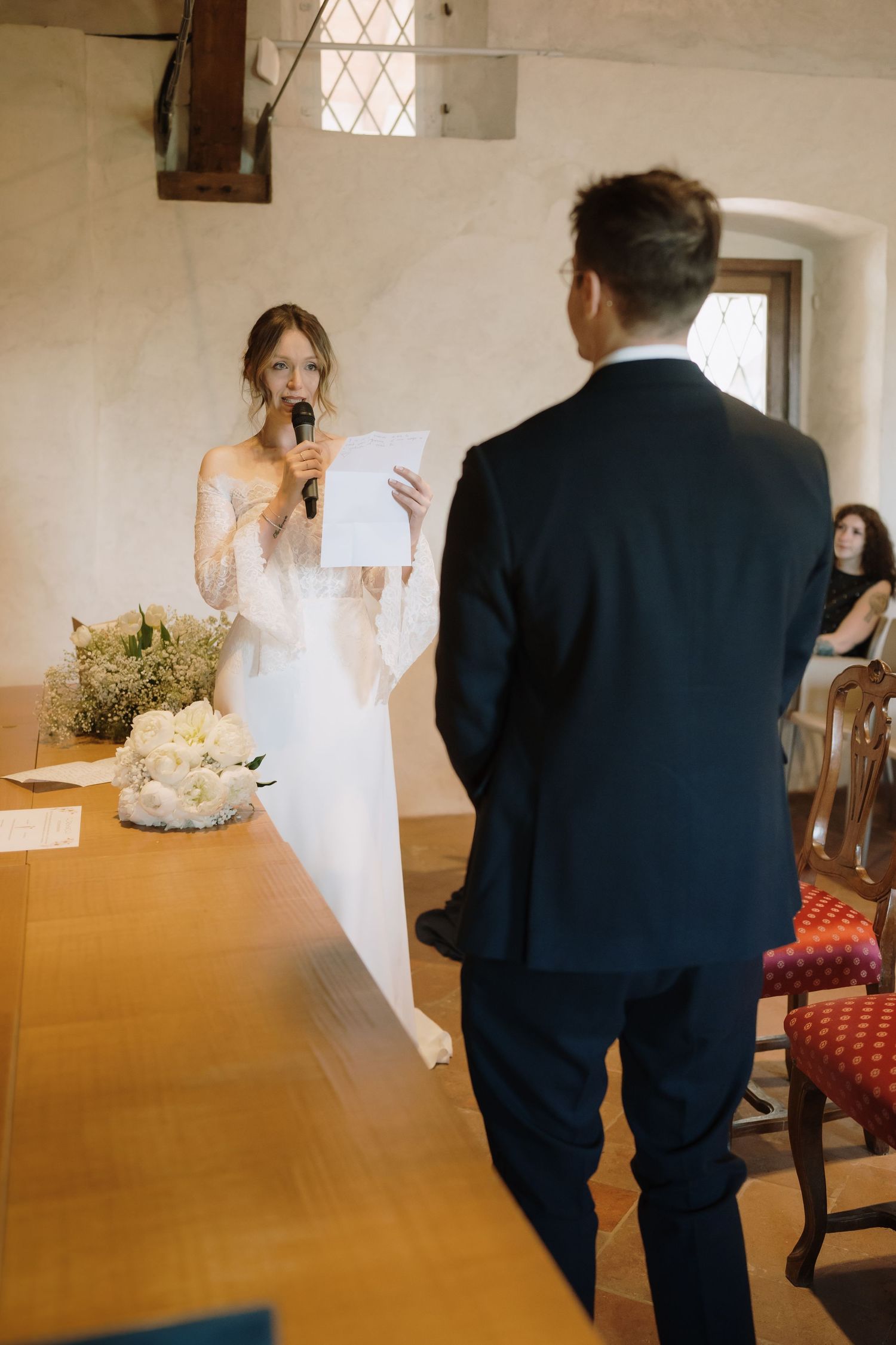 Bride in white lace gown reads wedding vows into microphone at altar while groom stands before her.